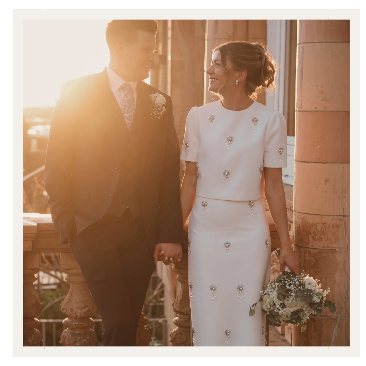 A bride and groom holding hands, smiling and looking at each other during sunset on a balcony, with warm sunlight and a cityscape in the background.