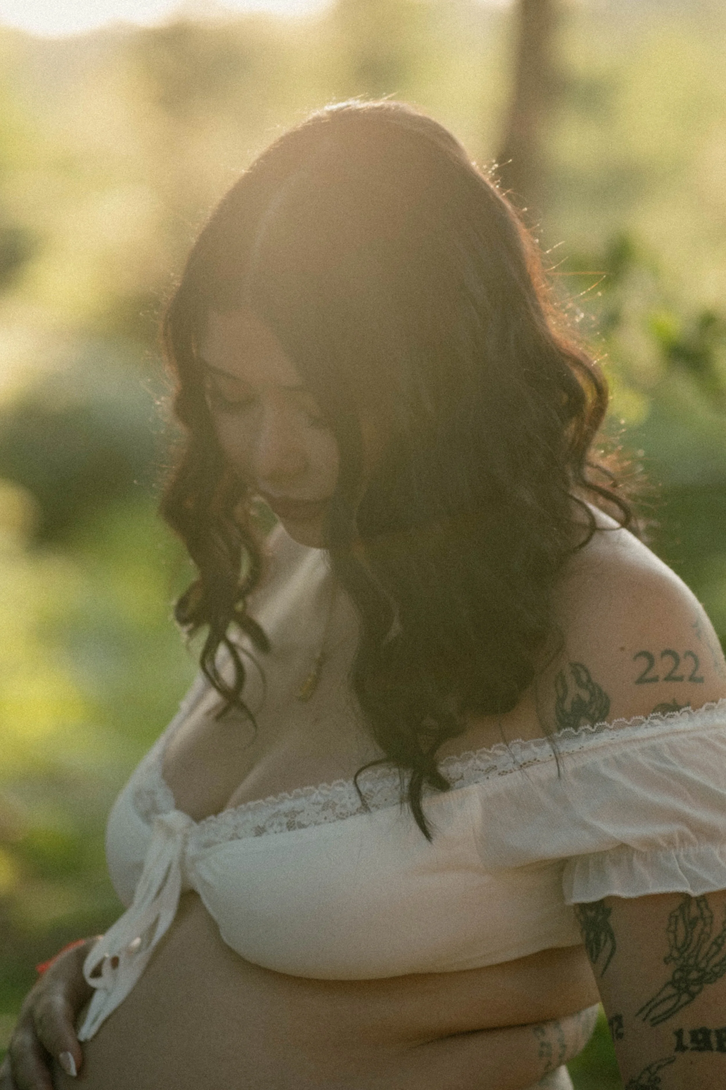 A woman with long, curly hair wearing a white off-shoulder top with lace details, tattooed arm, standing outdoors in a natural setting with sunlight and greenery.