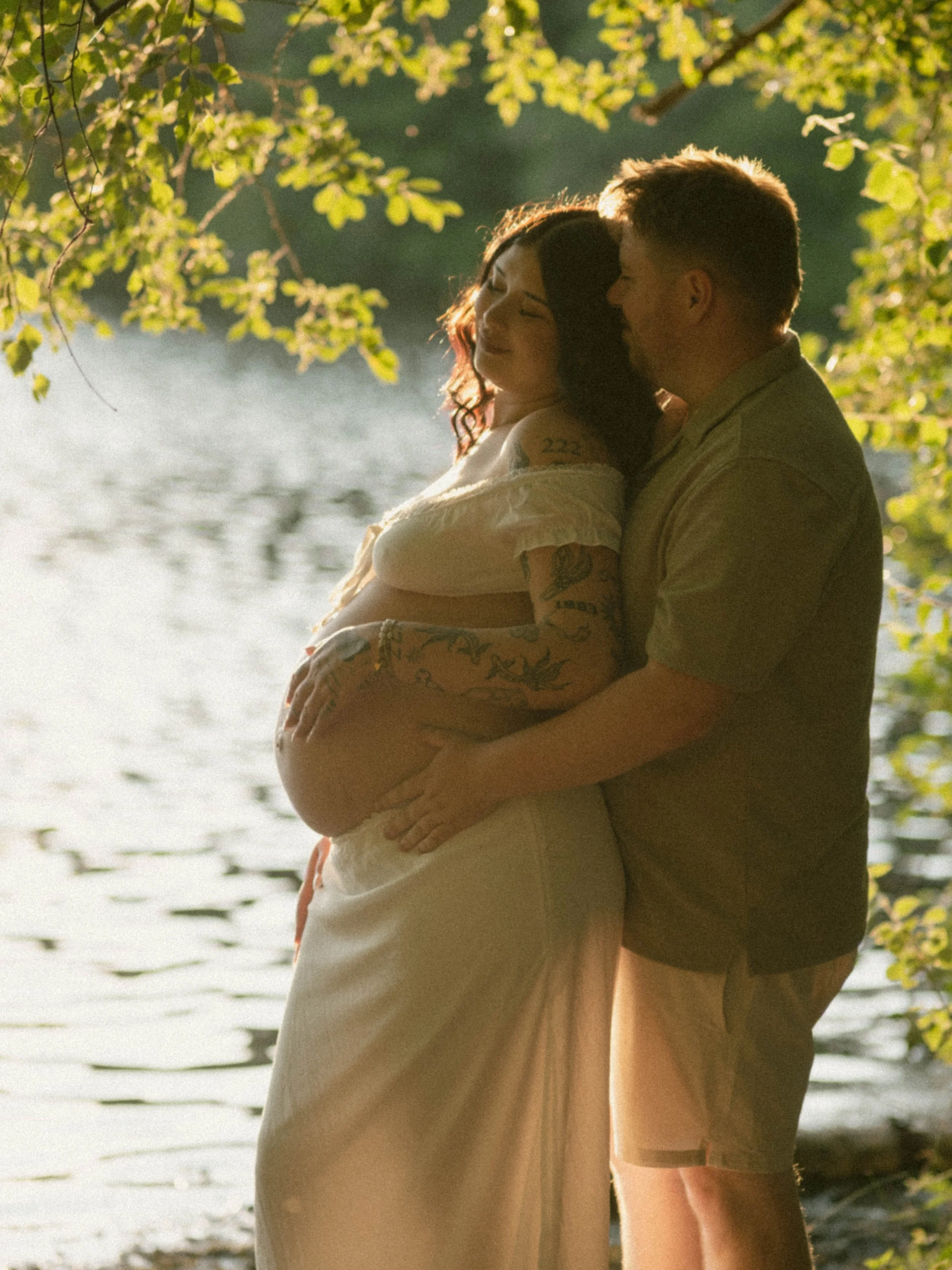 A couple embracing near a lake with green trees overhead and sunlight reflecting on the water.