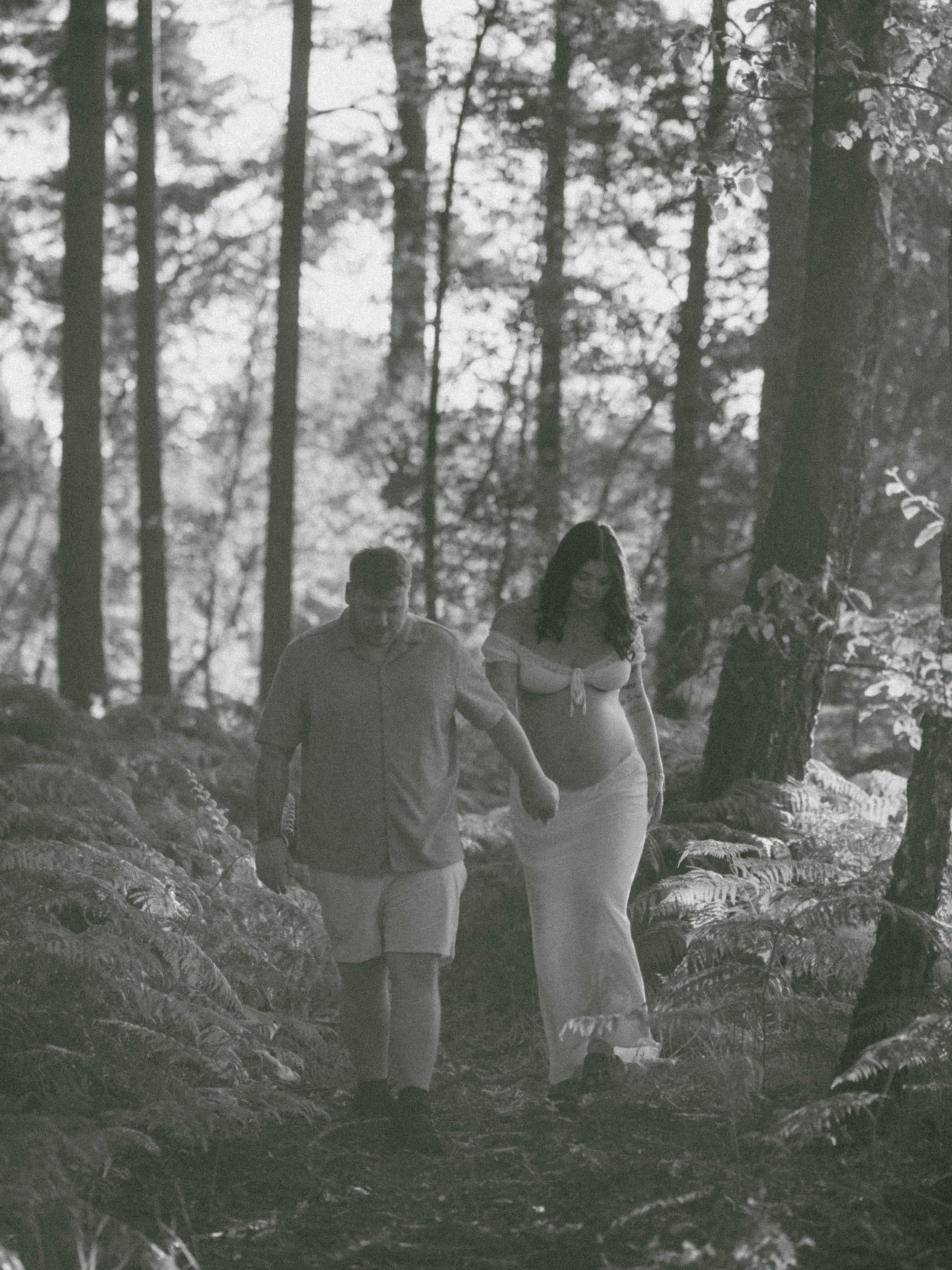 A man and a woman walking through a forest with tall trees and ferns, holding hands.