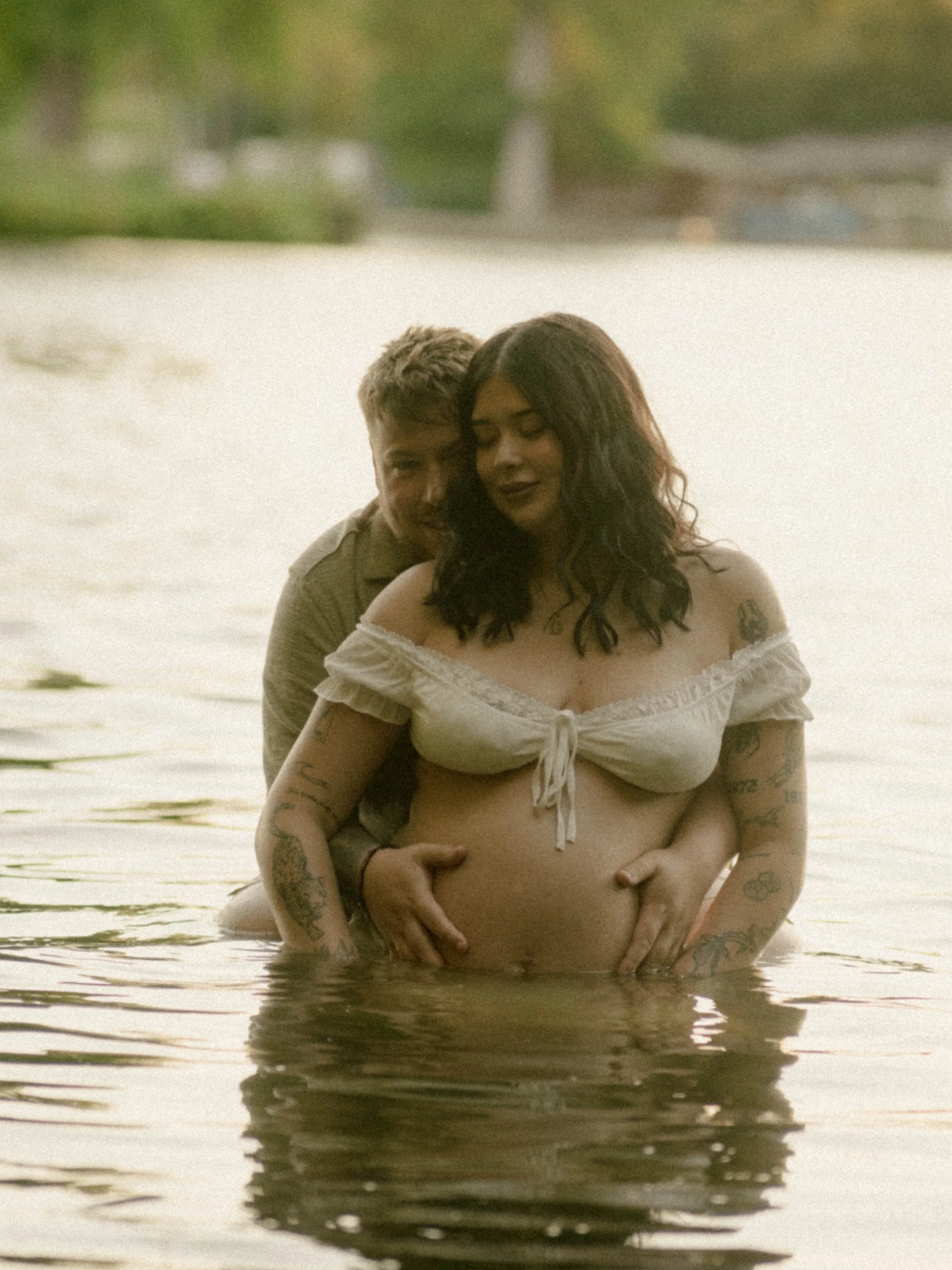 A couple is standing in shallow water at the beach, embracing each other. The woman, with dark wavy hair and tattoos on her arms, is wearing a white off-shoulder top, and the man, with short light hair and a tan shirt, is hugging her from behind. The