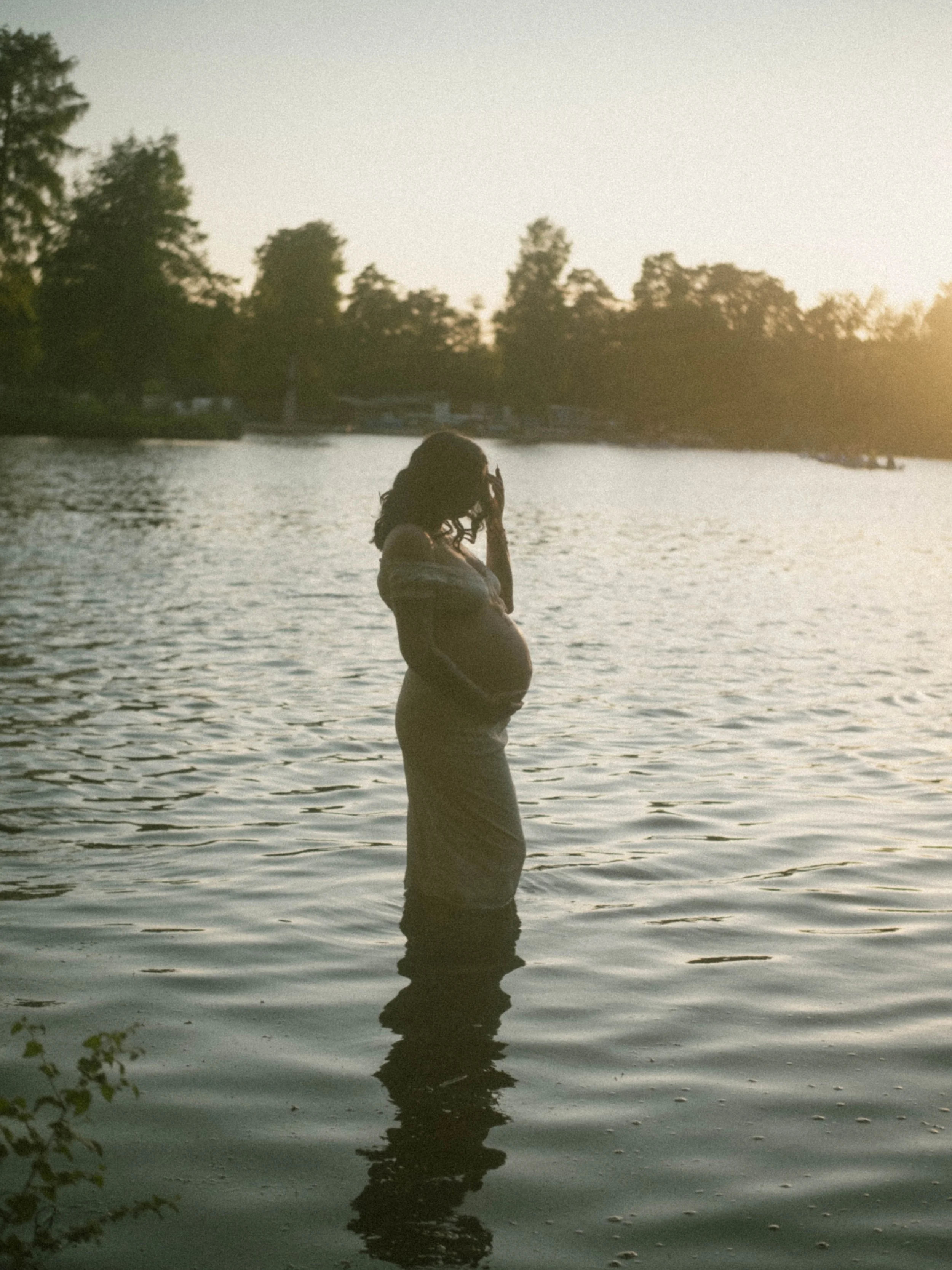 A pregnant woman standing in a body of water during sunset, with her hand near her face, and trees in the background.