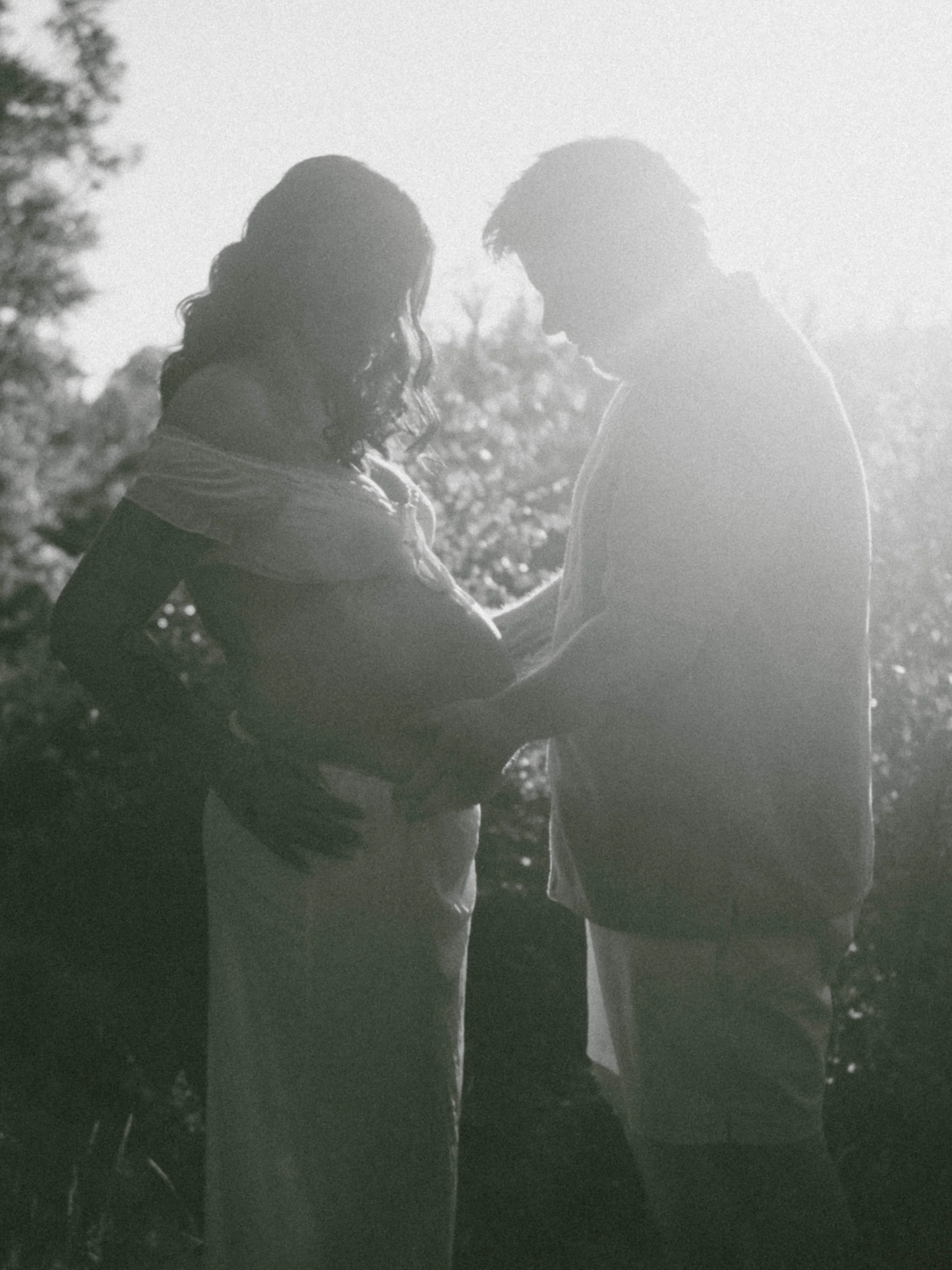 A black and white photo of a couple standing close, facing each other outdoors with trees in the background, sunlight coming through.