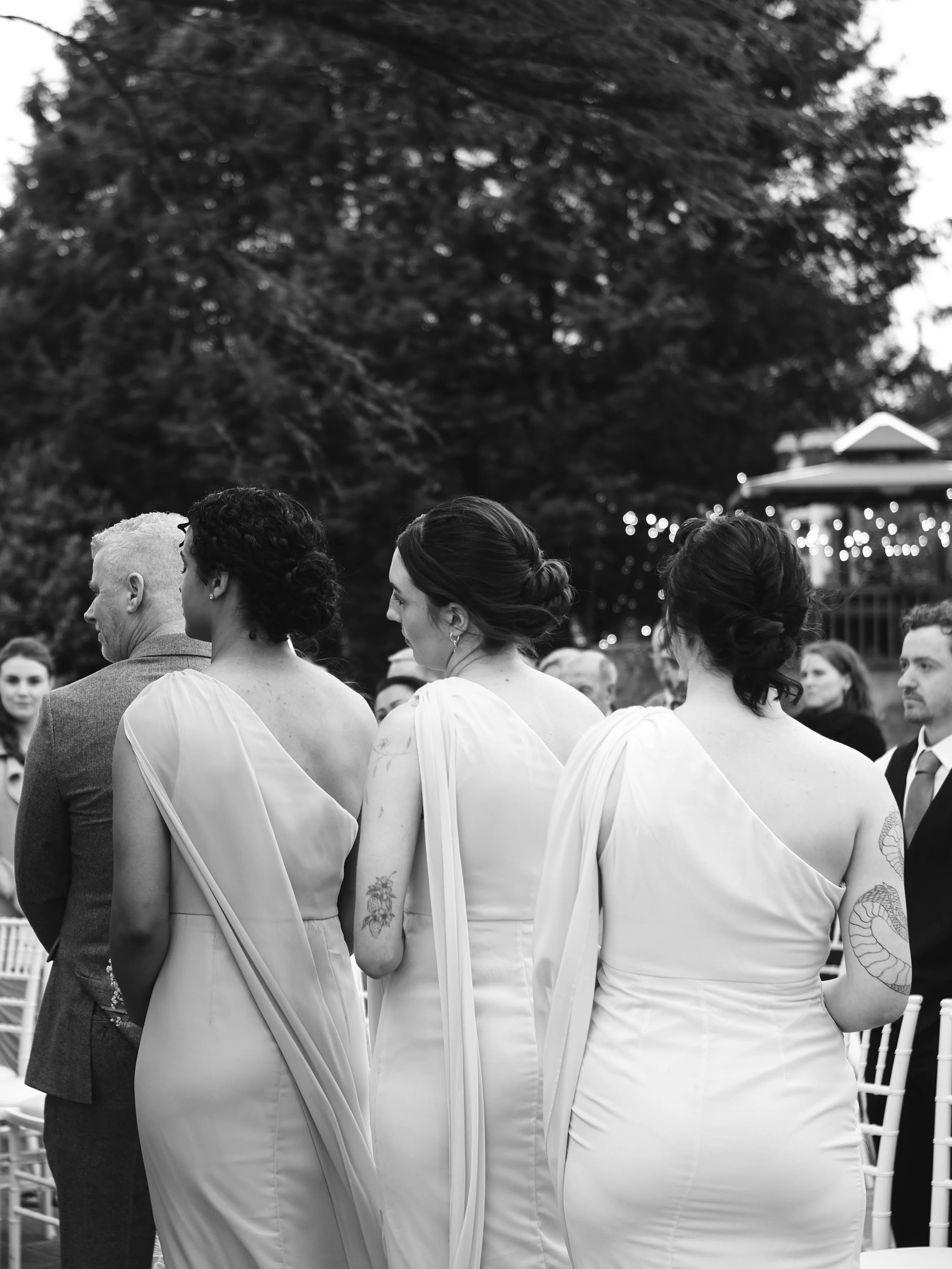 Black and white photo of several women in formal dresses standing outdoors, with a large tree and house in the background at dusk.