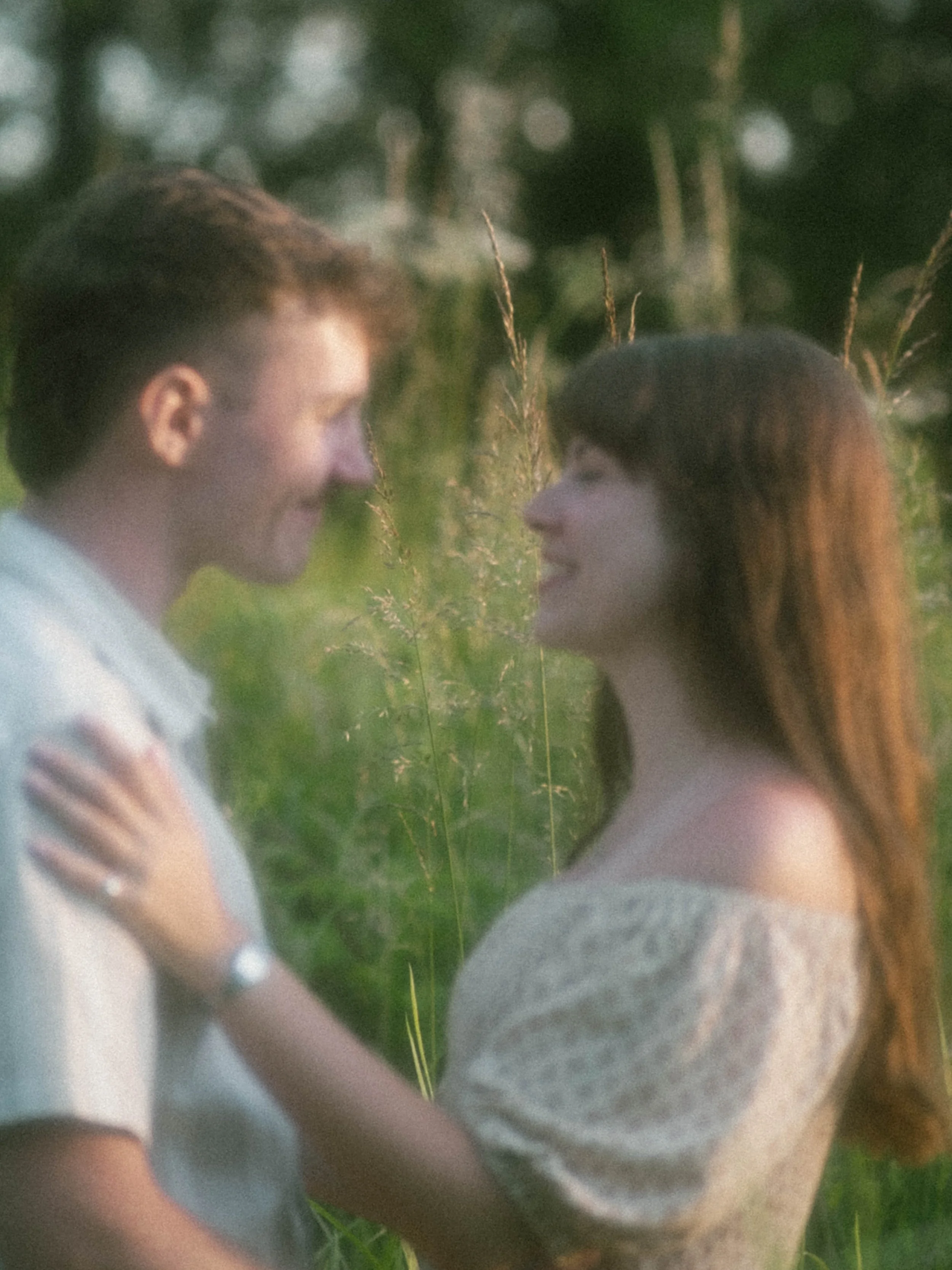 A woman with brown hair and a man with brown hair are facing each other outdoors, holding a large green leaf between them.