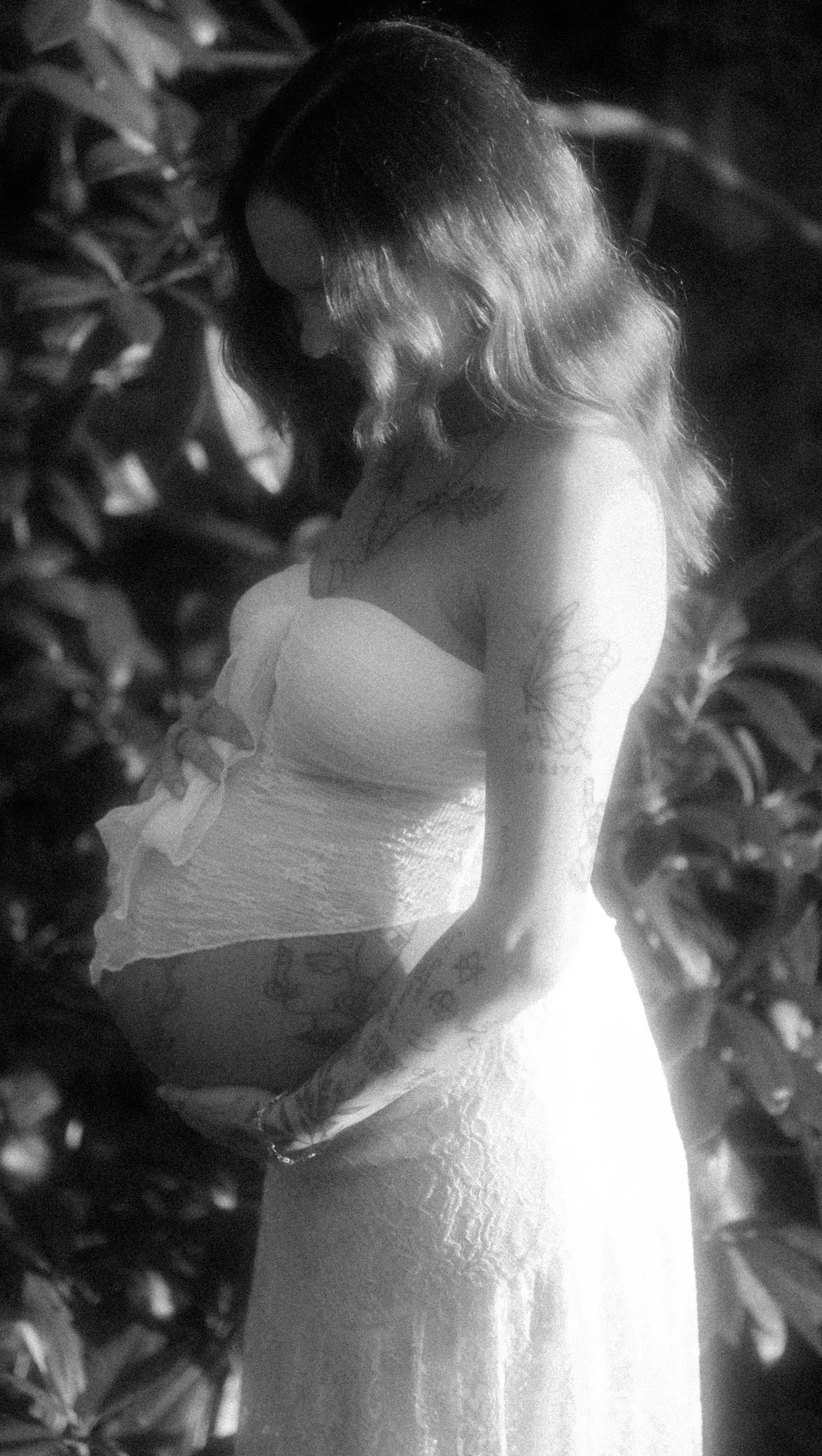 A black and white photo of a woman with shoulder-length hair, wearing a strapless dress, surrounded by foliage, standing by a body of water with sunlight reflecting on the surface.