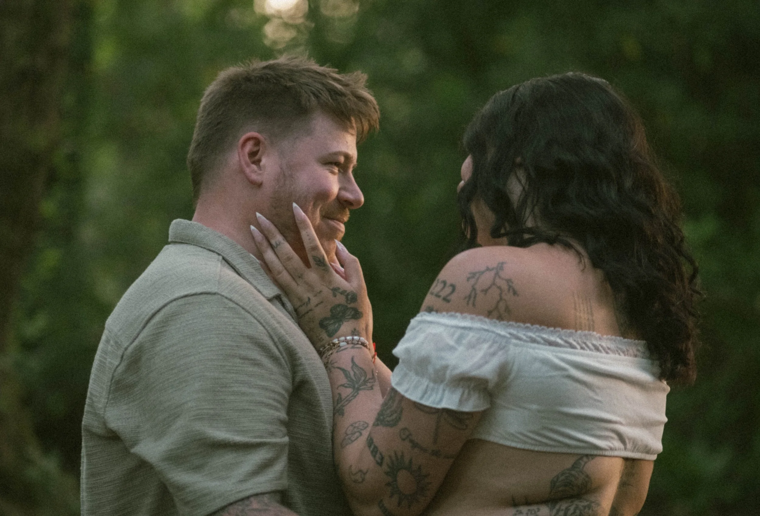 A young man and woman are sharing an intimate moment outdoors, looking into each other's eyes with smiles. They are close, with the woman touching the man's face gently, and both are standing against a background of greenery.