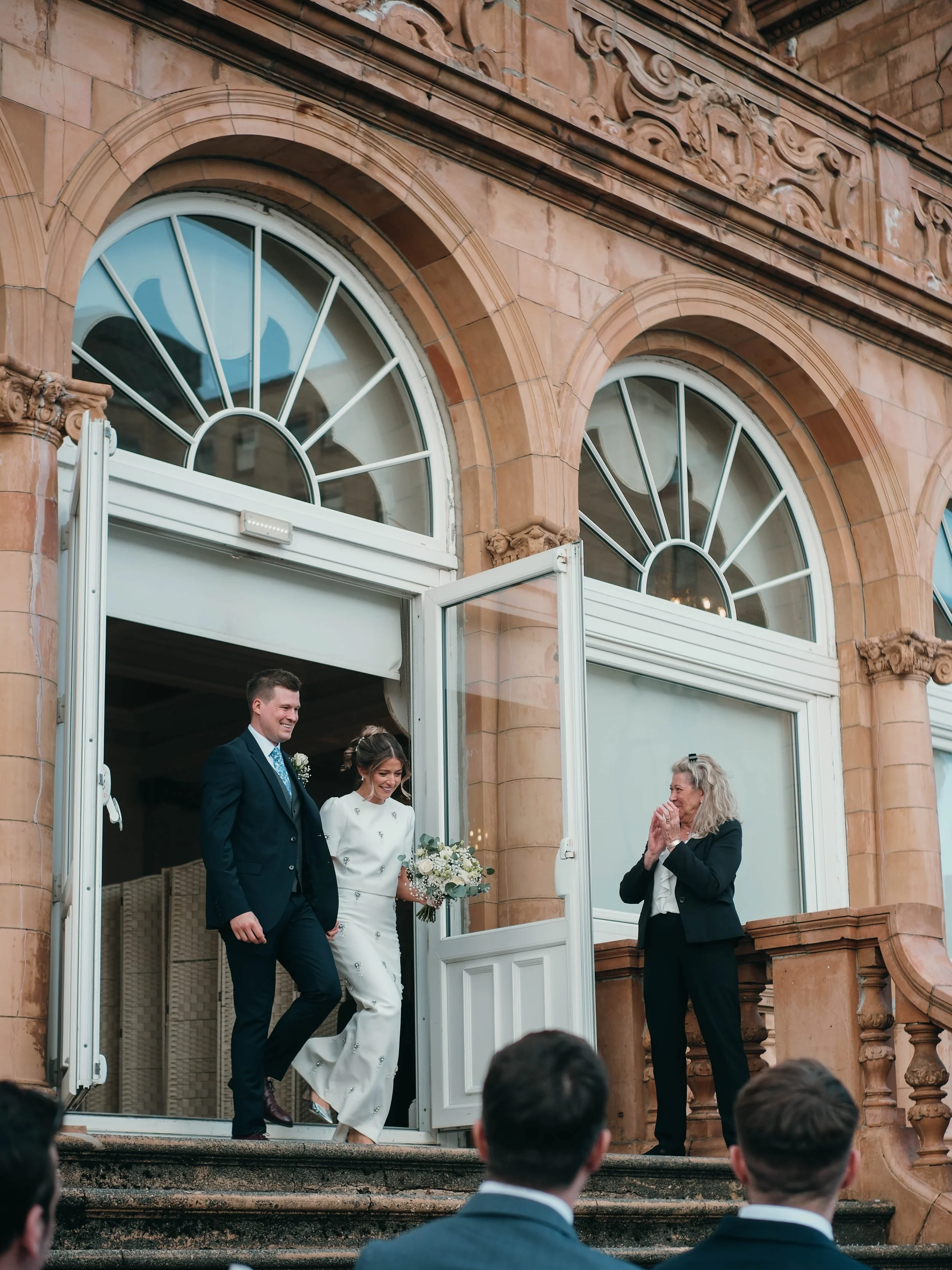 A wedding ceremony taking place on the steps of a historic building with ornate stonework. The bride and groom are just inside the doorway, smiling and holding hands, with a woman standing nearby. The groom is in a dark suit with a blue tie, and the 