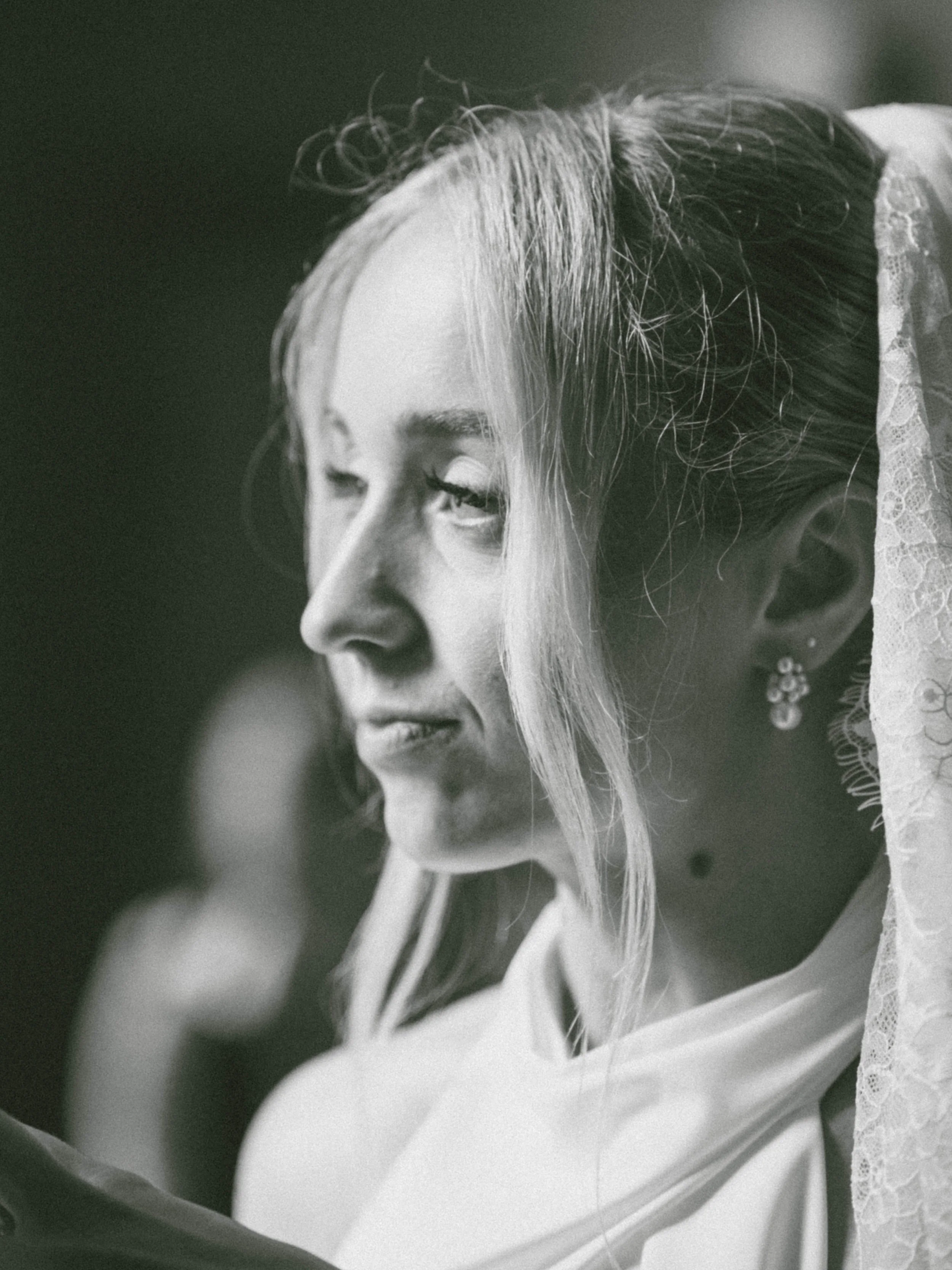 A close-up black and white photograph of a woman with light-colored hair, earrings, and a lace garment, looking downward with a serene expression.