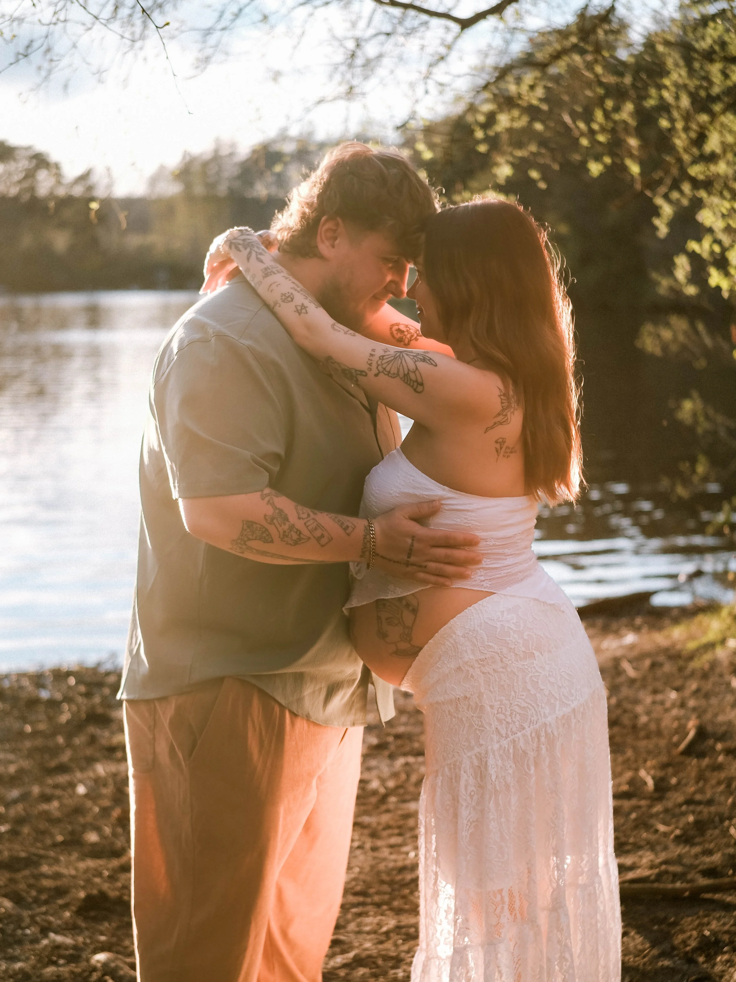 A couple is embracing by a lake at sunset, with trees in the background, sharing a close moment.
