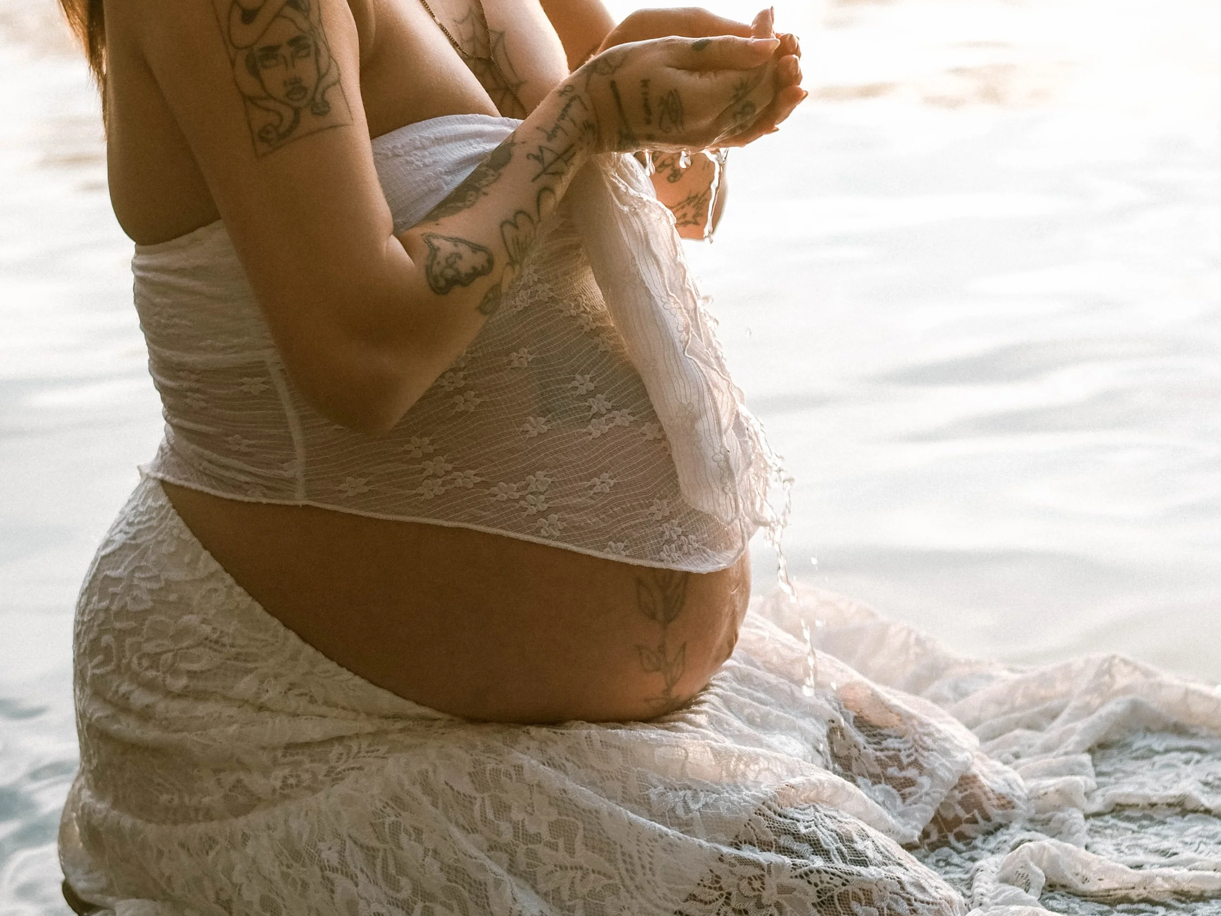 Close-up of a person with tattoos on their arm, holding water with both hands, wearing a light lace dress, sitting near water during sunset or sunrise.