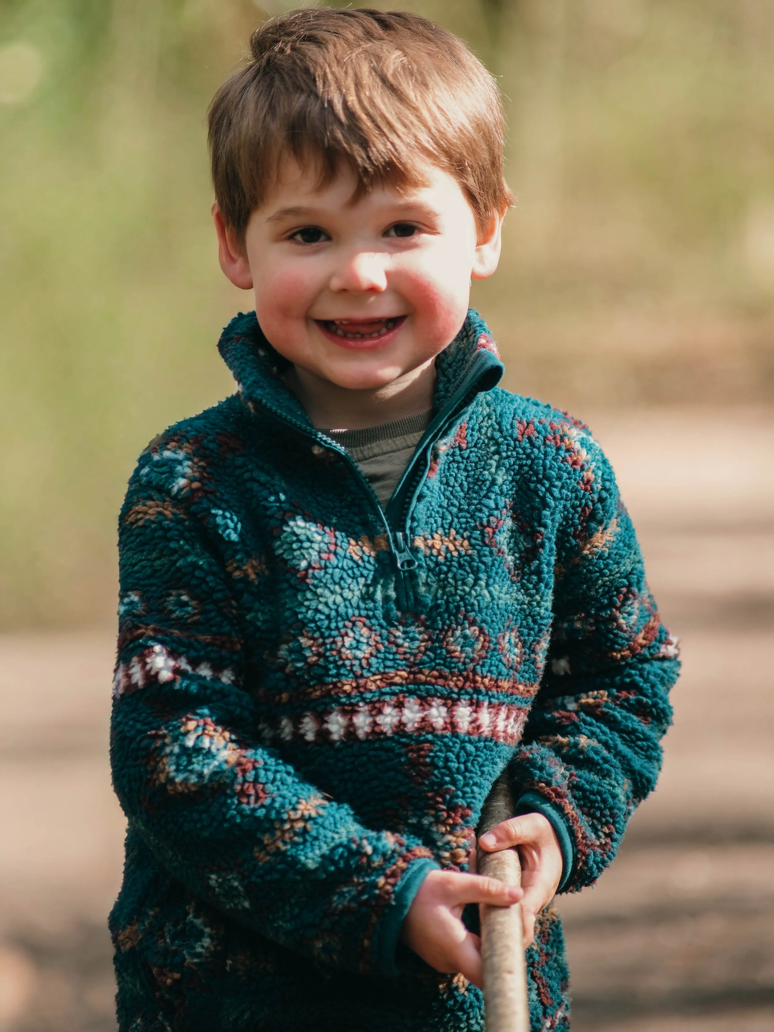 A young boy with brown hair and rosy cheeks smiling outdoors, wearing a multicolored fleece jacket, holding a stick.