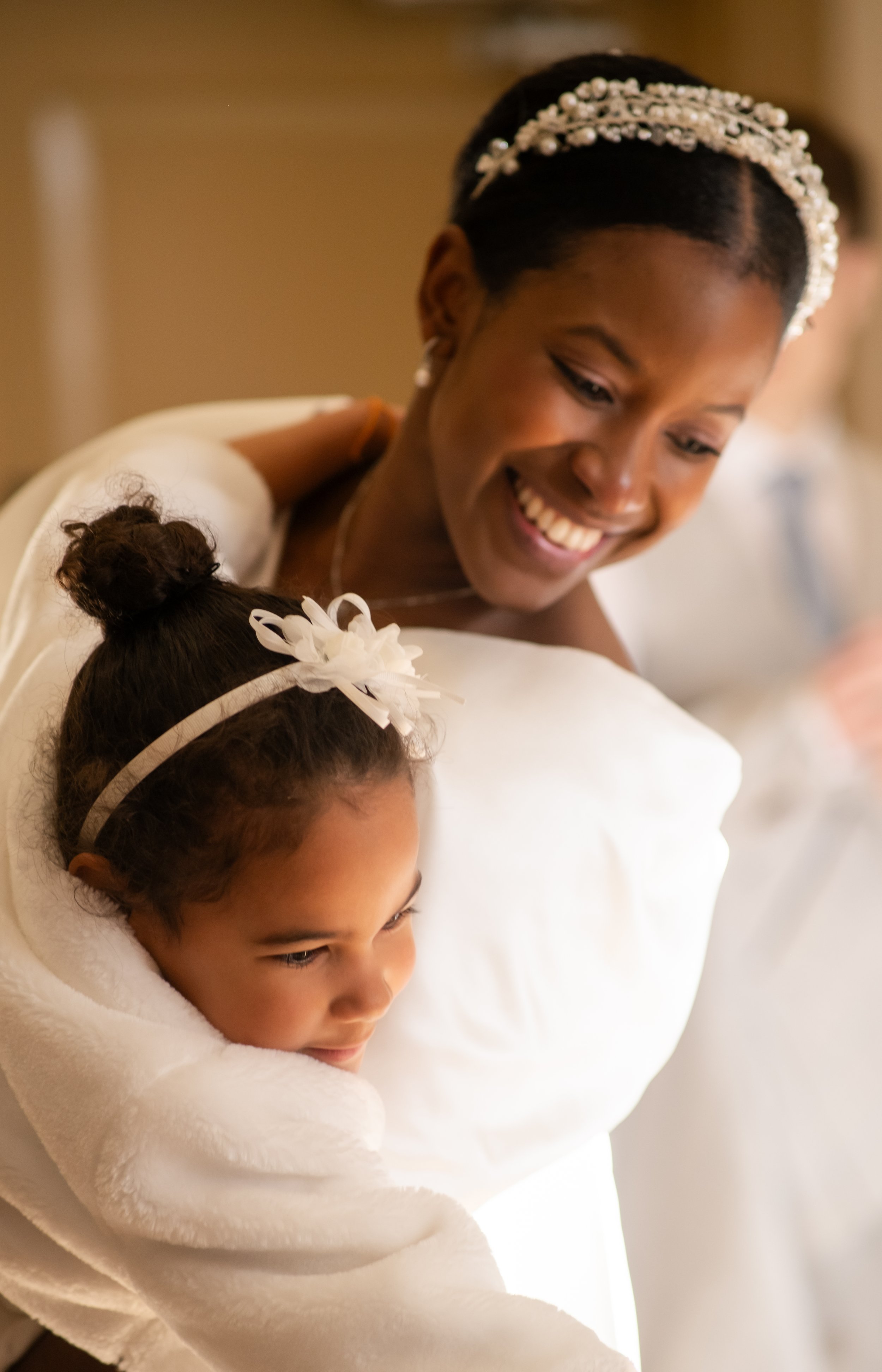 A woman in a white dress and pearl headband smiles as she looks down at a young girl with a bun hairstyle and white flower hair accessory, who is wrapped in a white blanket.