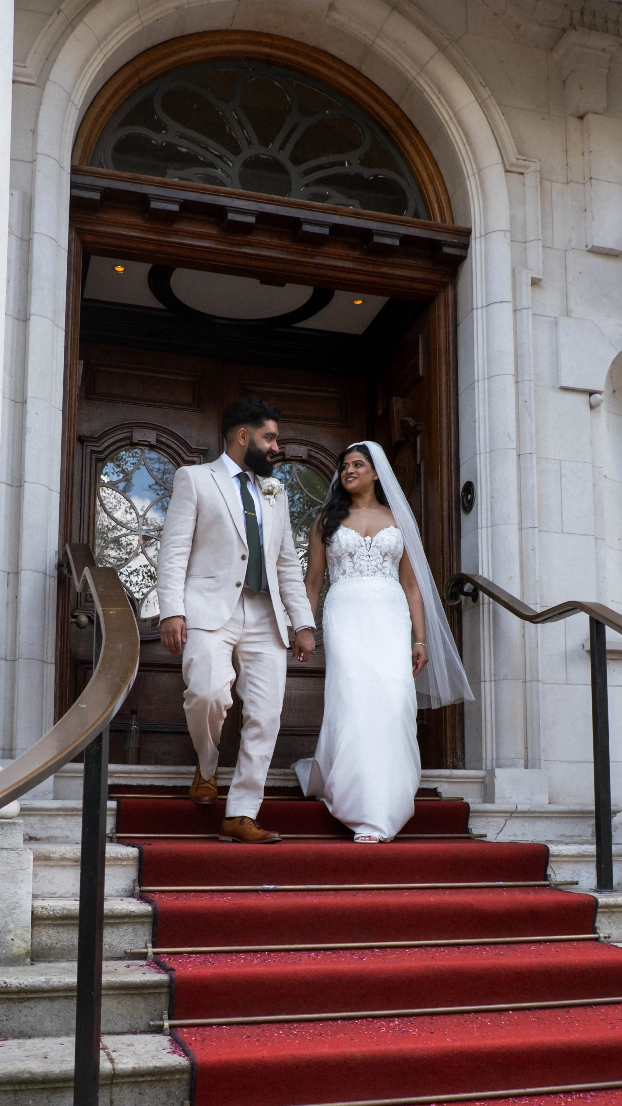 A bride and groom holding hands and smiling as they walk down the steps of a church with a red carpet. The bride is wearing a white wedding gown with a veil, and the groom is wearing a light-colored suit with a dark tie.