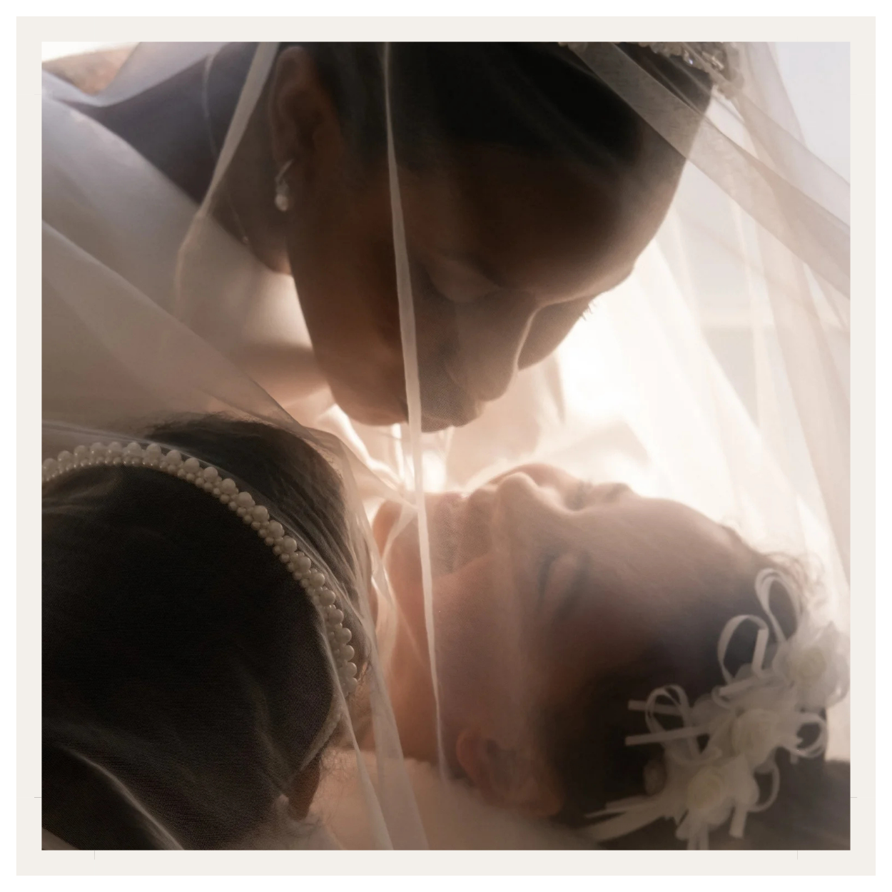 A couple of women lying under a sheer curtain, touching foreheads and smiling, with one wearing a pearl headband and the other with curlers in her hair, in a close, intimate moment.