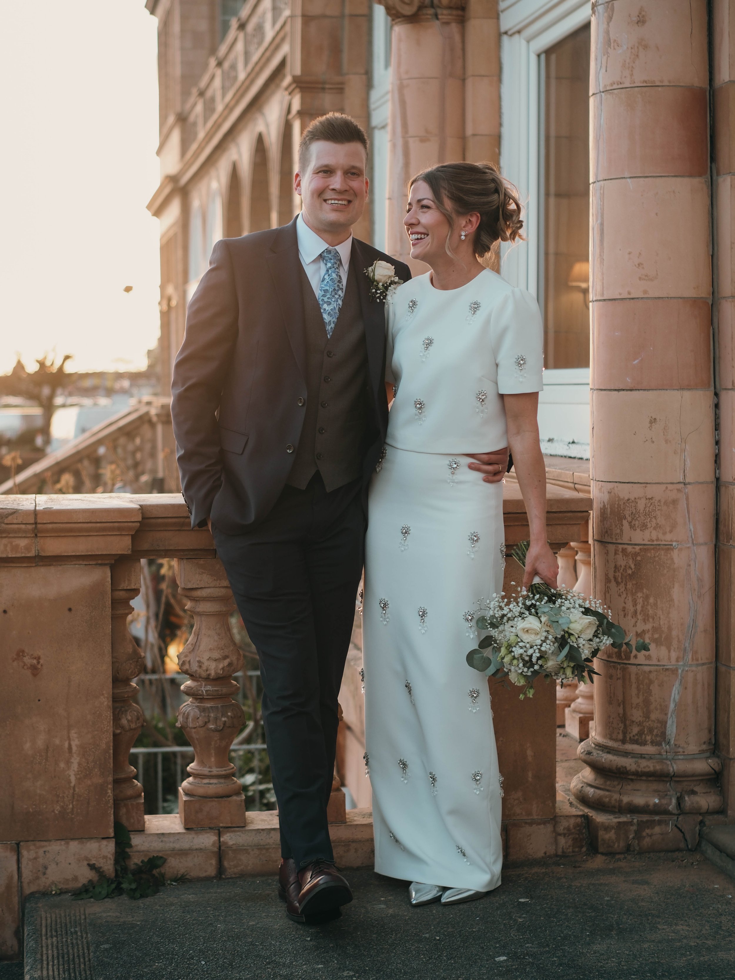 A smiling groom and bride stand together on a balcony, dressed in wedding attire, with the groom in a dark suit and the bride in a white gown holding a bouquet of flowers, in front of a historic building.