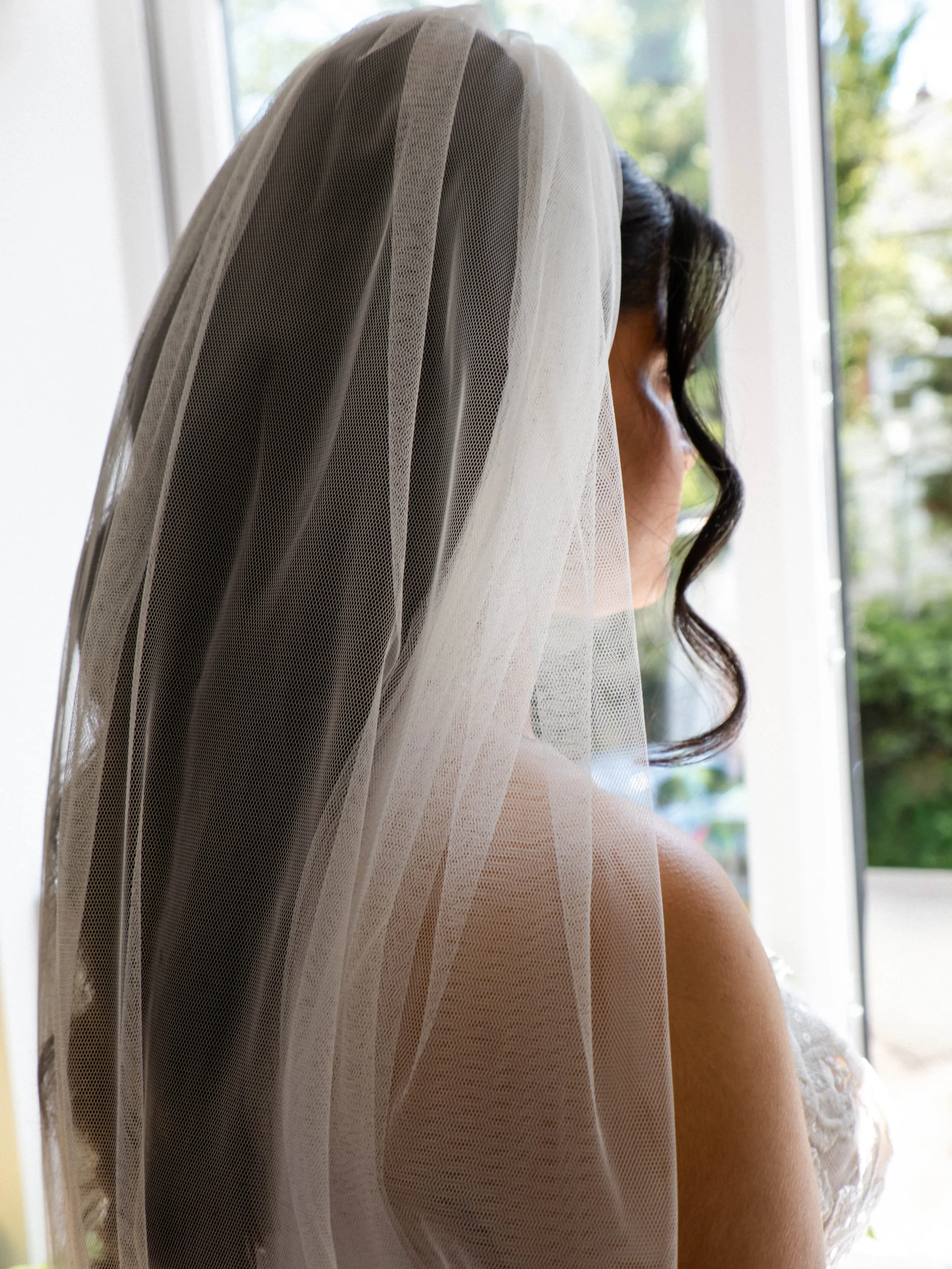 A woman with dark wavy hair looking out a window, wearing a white, sheer veil with lace details on the sleeves.