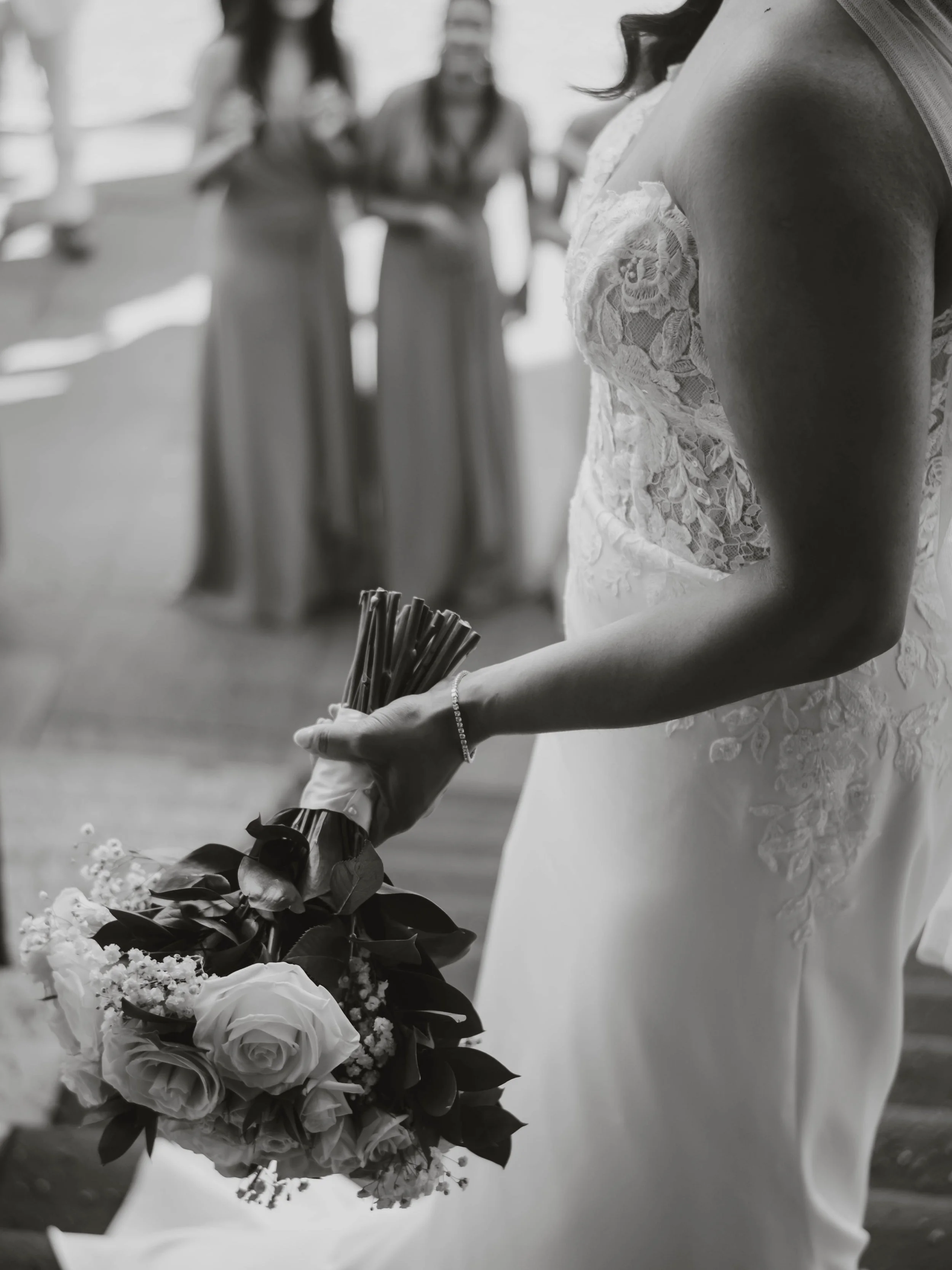 Close-up of a bride in a lace wedding dress holding a bouquet of roses and greenery during a wedding ceremony, with two women in dresses in the background.