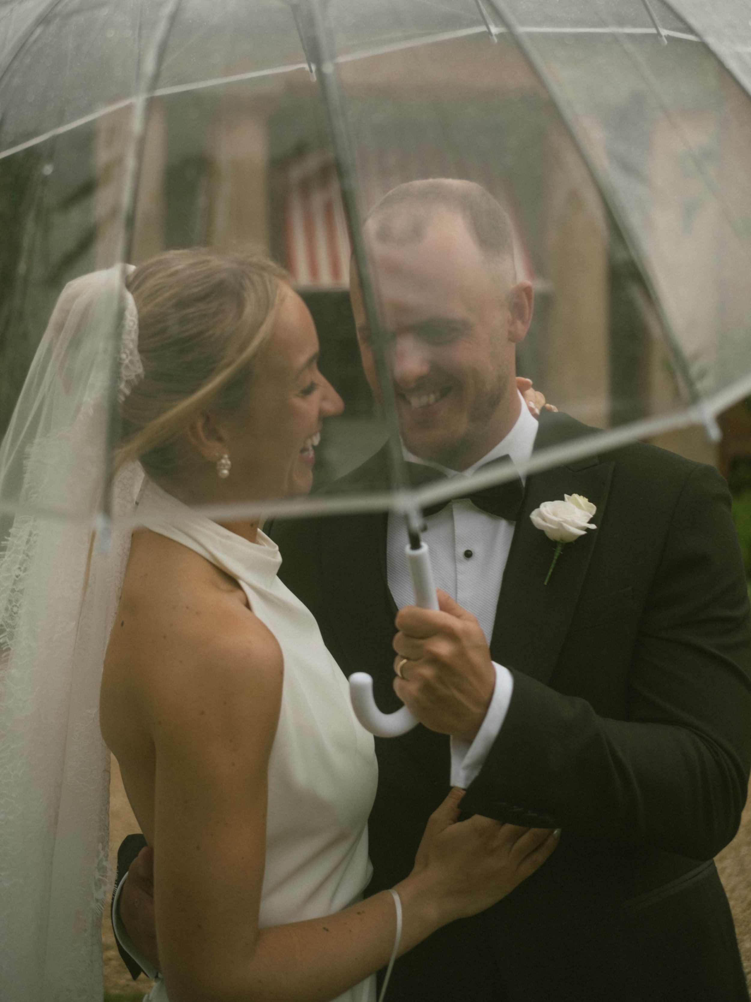A bride and groom sharing a kiss behind a clear umbrella, with the bride wearing a white dress and veil, and the groom in a black tuxedo with a white boutonnière, during a wedding celebration.