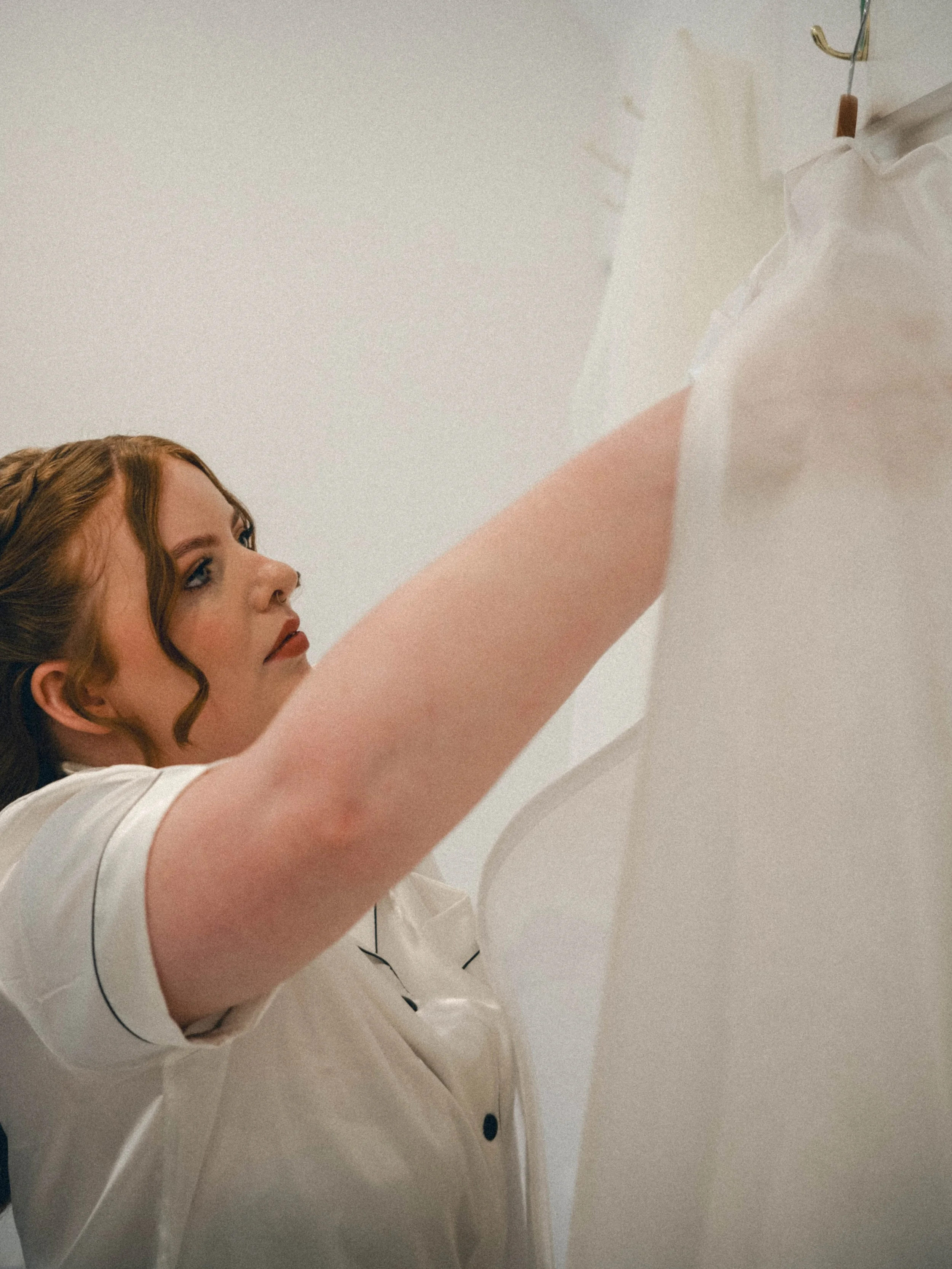 A woman with red hair styled in curls, wearing a white uniform, is reaching upward to hang or adjust something on a hook attached to the wall.