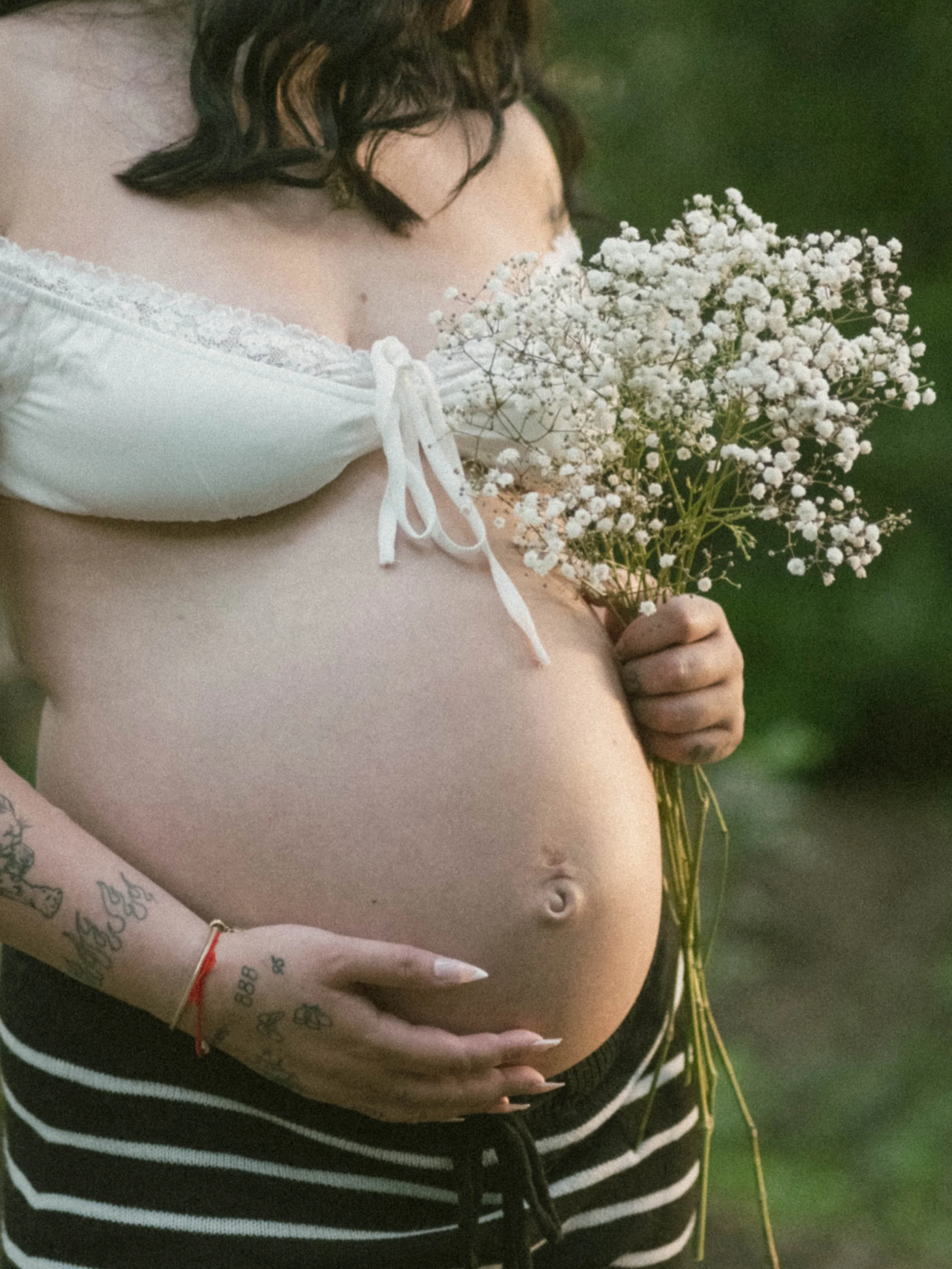 Person with tattoos holding a bunch of white baby's breath flowers, wearing a white off-shoulder top with a bow tie, and black and white striped pants.