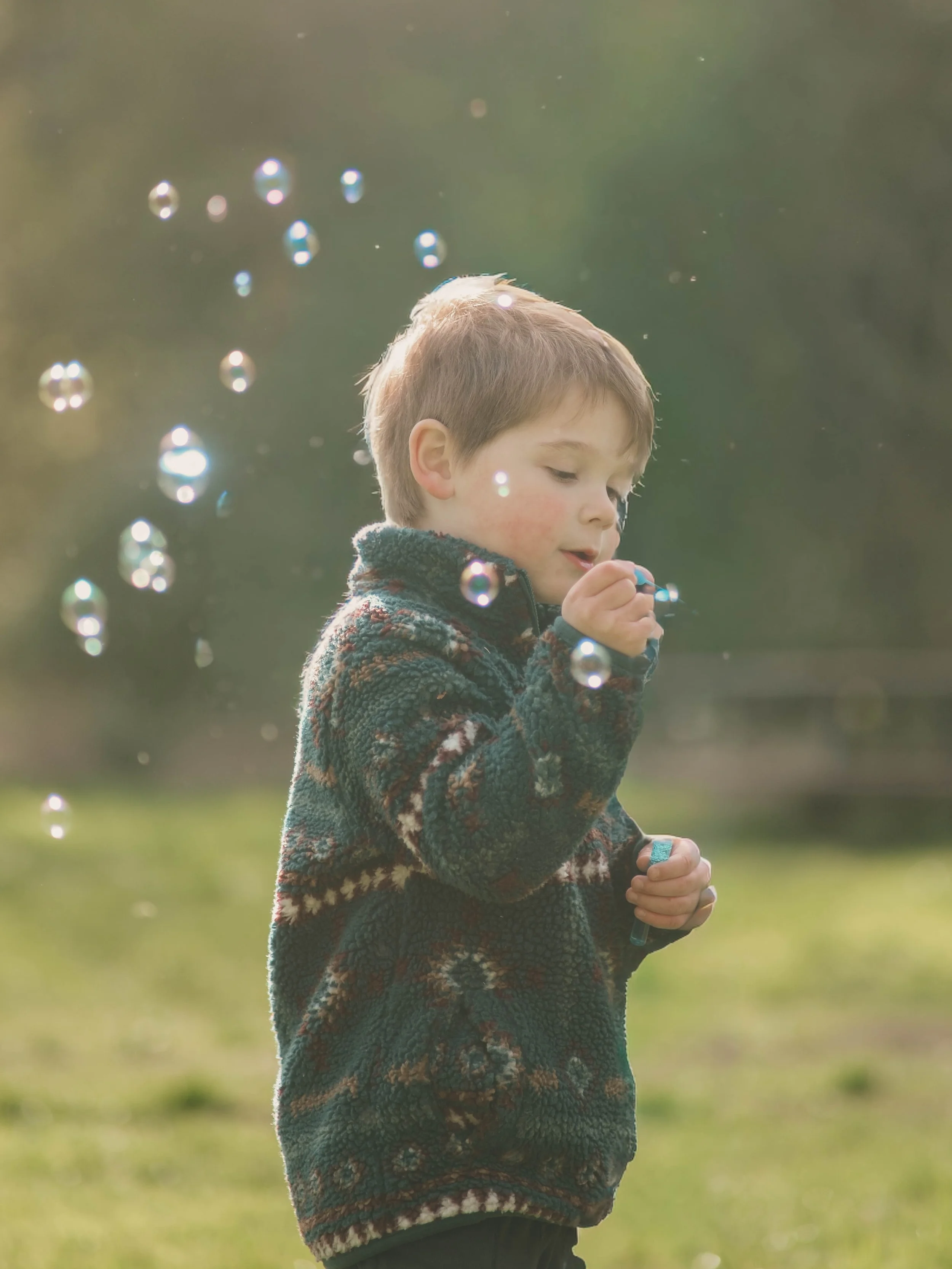 Young boy outdoors in a fleece sweater with a pattern, holding a bubble wand, surrounded by floating bubbles, with a blurred green background.