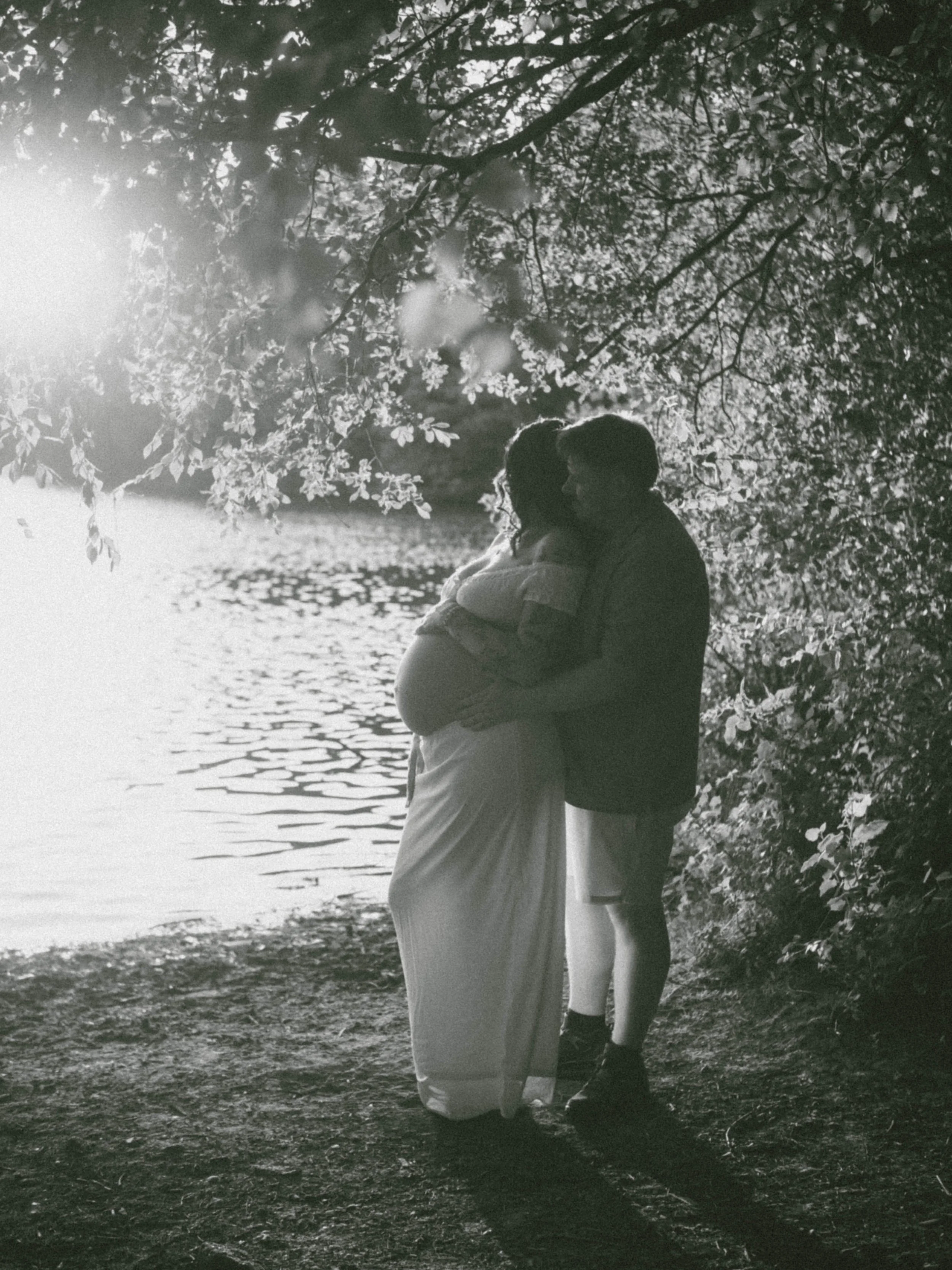 A silhouette of a pregnant woman and a young girl standing beneath a tree near a body of water at sunset.