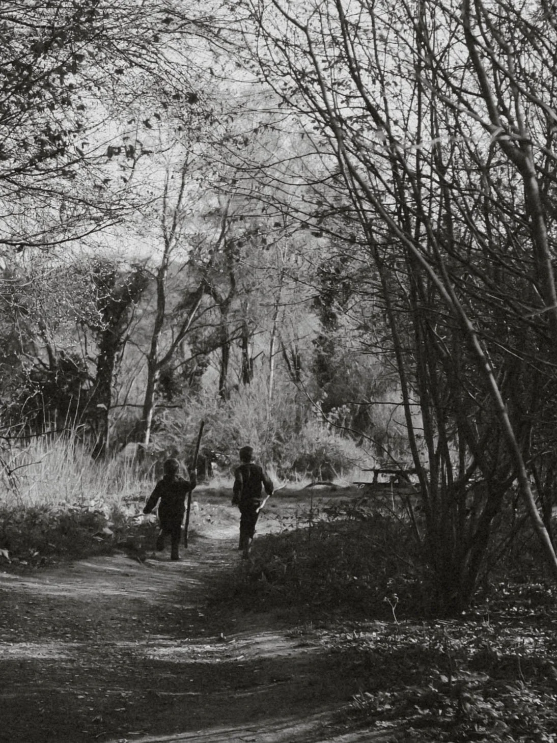 Two children walking along a dirt trail in a wooded area with leafless trees and shrubs, black and white photograph.
