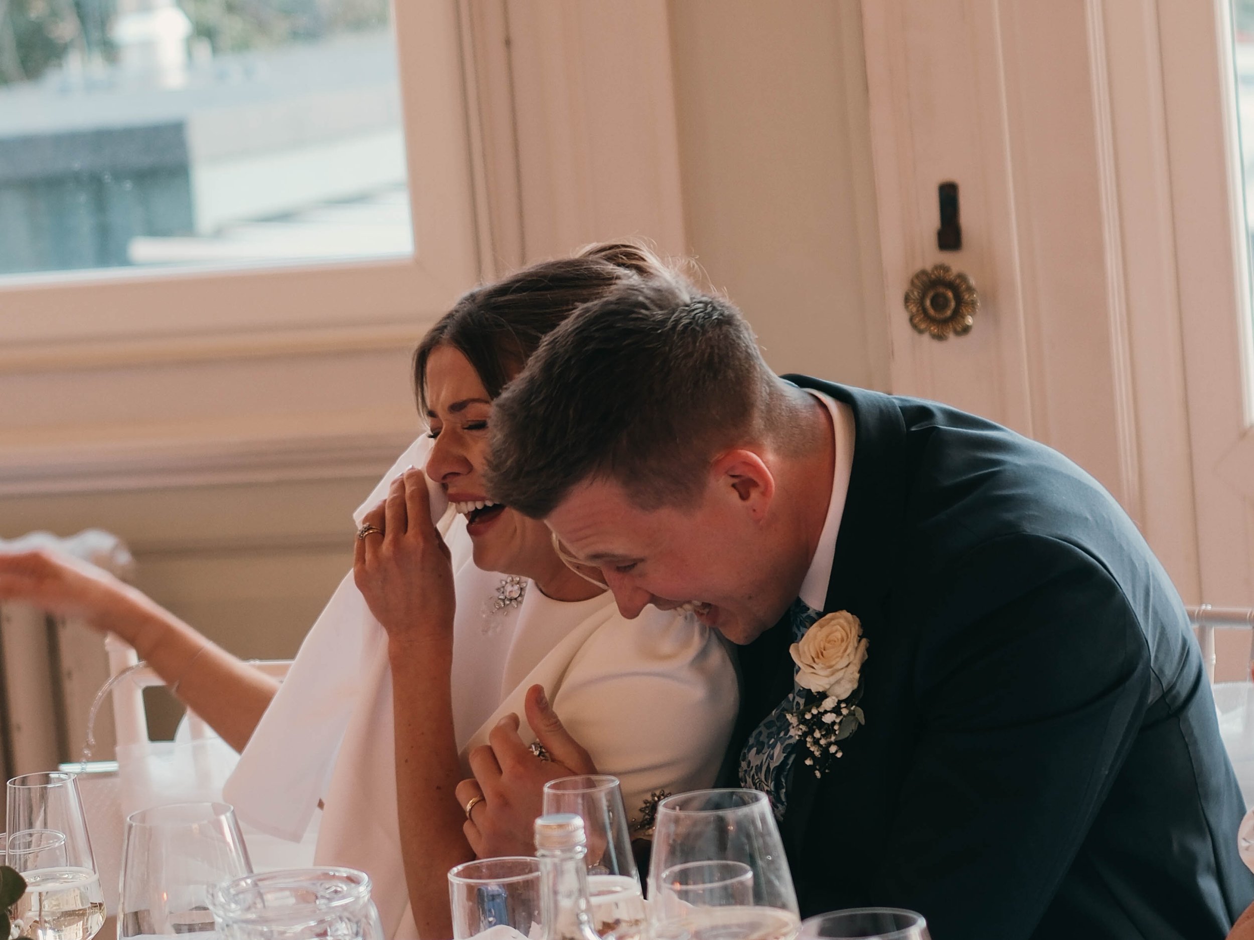 A newlywed couple sharing an emotional moment at their wedding reception, sitting at a table with glasses and bottles around them, inside a room with large windows.