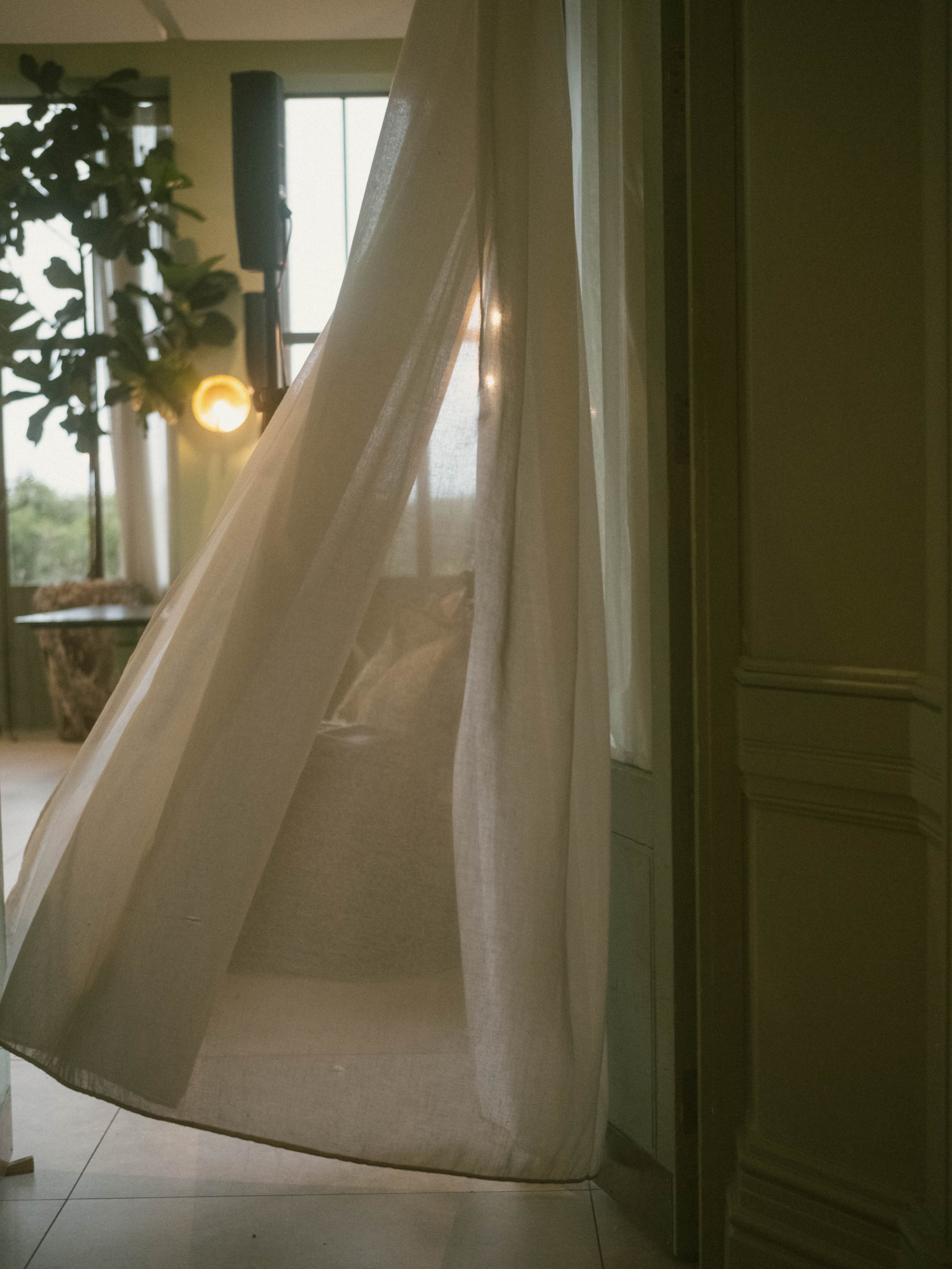 A bedroom with sheer curtains, a window, a potted plant, and a standing lamp in the background.