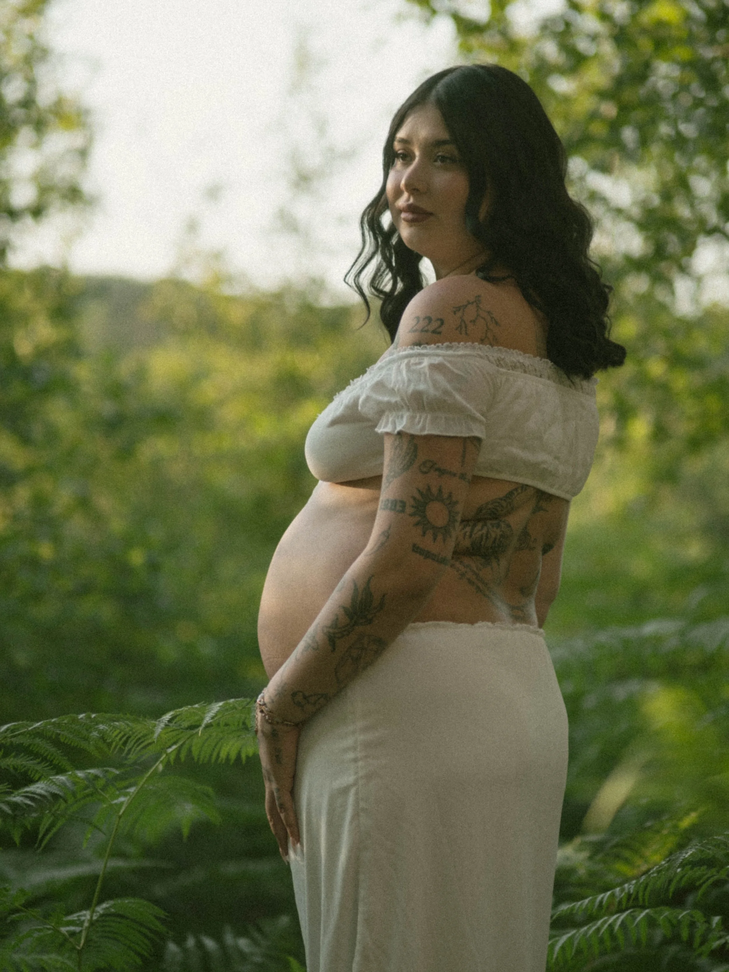 A woman with black wavy hair and tattoos on her arm and shoulder, wearing a white off-shoulder top and skirt, standing outdoors surrounded by green foliage and looking into the distance.