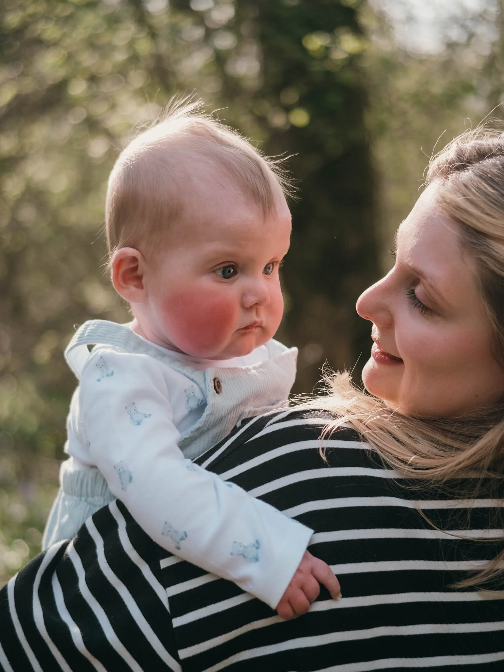 A woman with blonde hair holding a young child with blonde hair and blue eyes outdoors with blurred trees in the background.