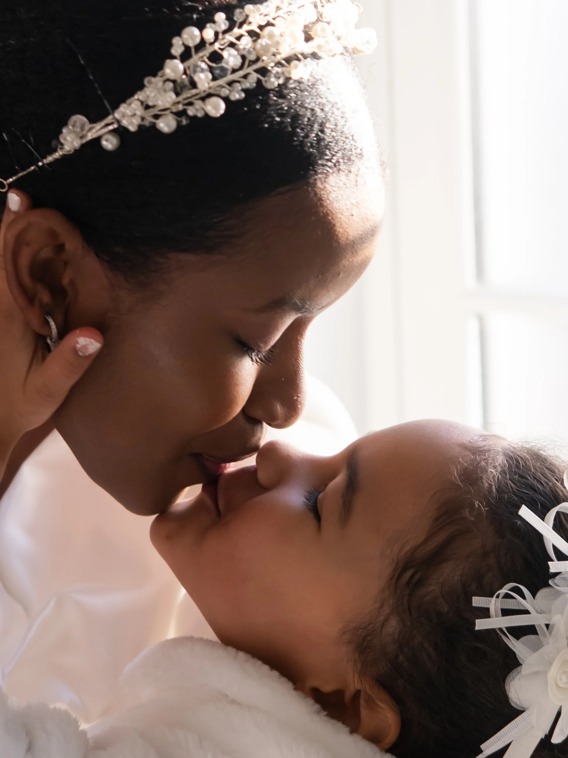 A woman with a pearl hair accessory and earrings tenderly kisses a baby, both with eyes closed, in a close-up intimate moment.