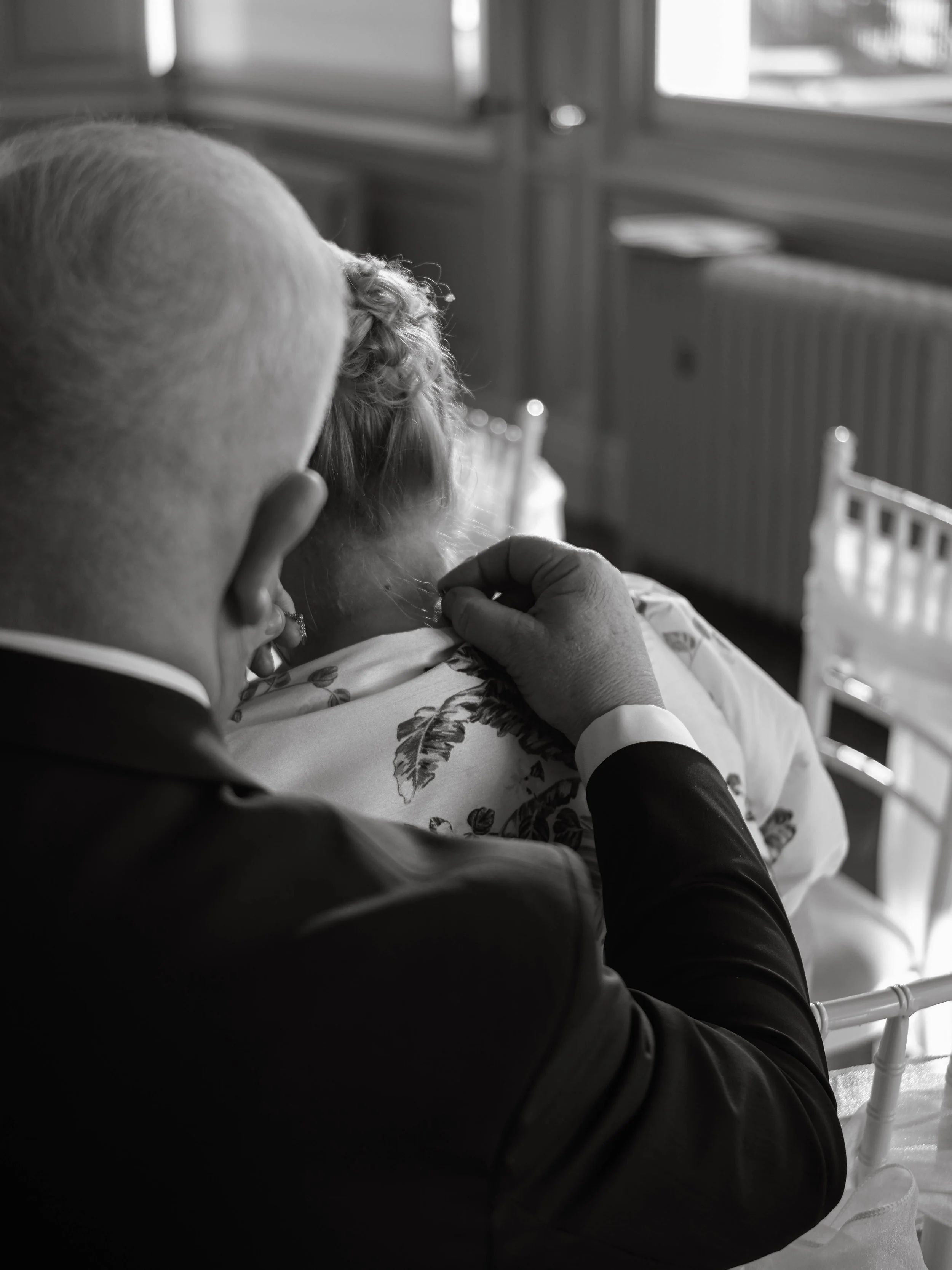A person with short hair and earrings is sitting near a window, with another person, possibly a caregiver, gently touching their shoulder. The setting appears to be a hospital or care facility, indicated by a bed and medical equipment nearby.