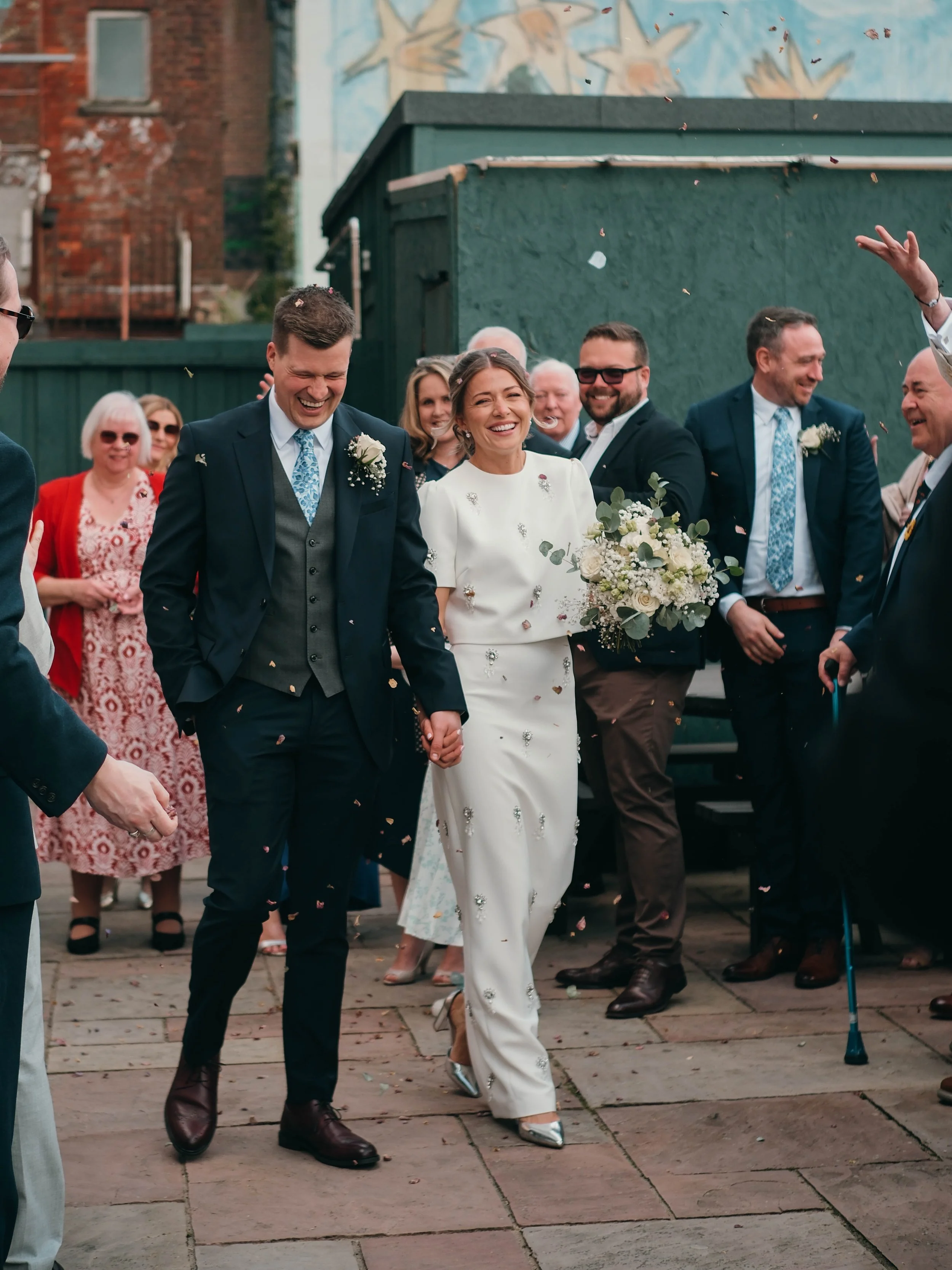 A group of people, including a bride in a white dress and a groom in a dark suit, are smiling and celebrating outdoors. The bride is holding a bouquet, and everyone is throwing confetti.