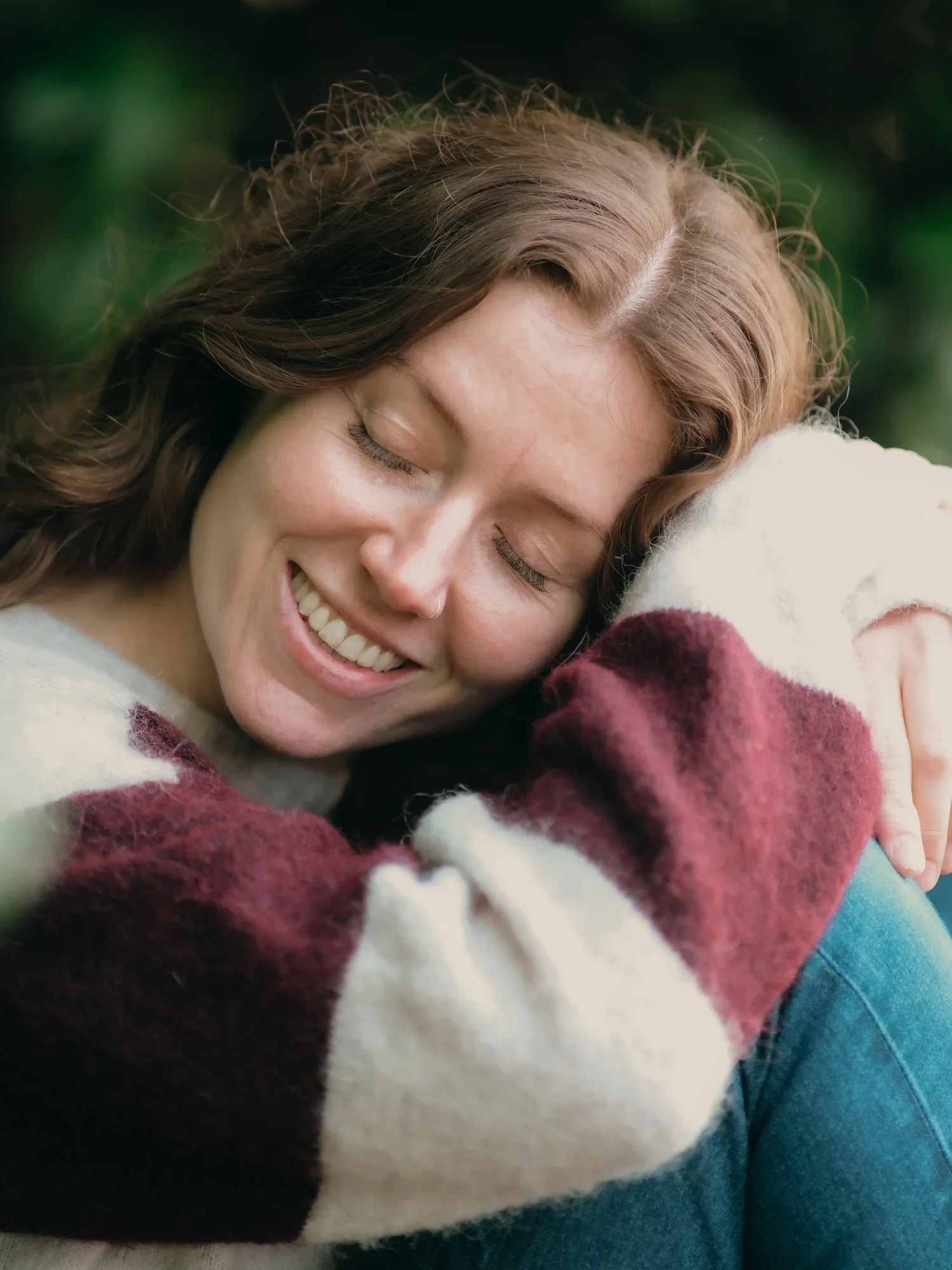 A woman with light skin and curly brown hair smiling with her eyes closed as she hugs a dog with a multicolored sweater, outdoors with green blurred background.