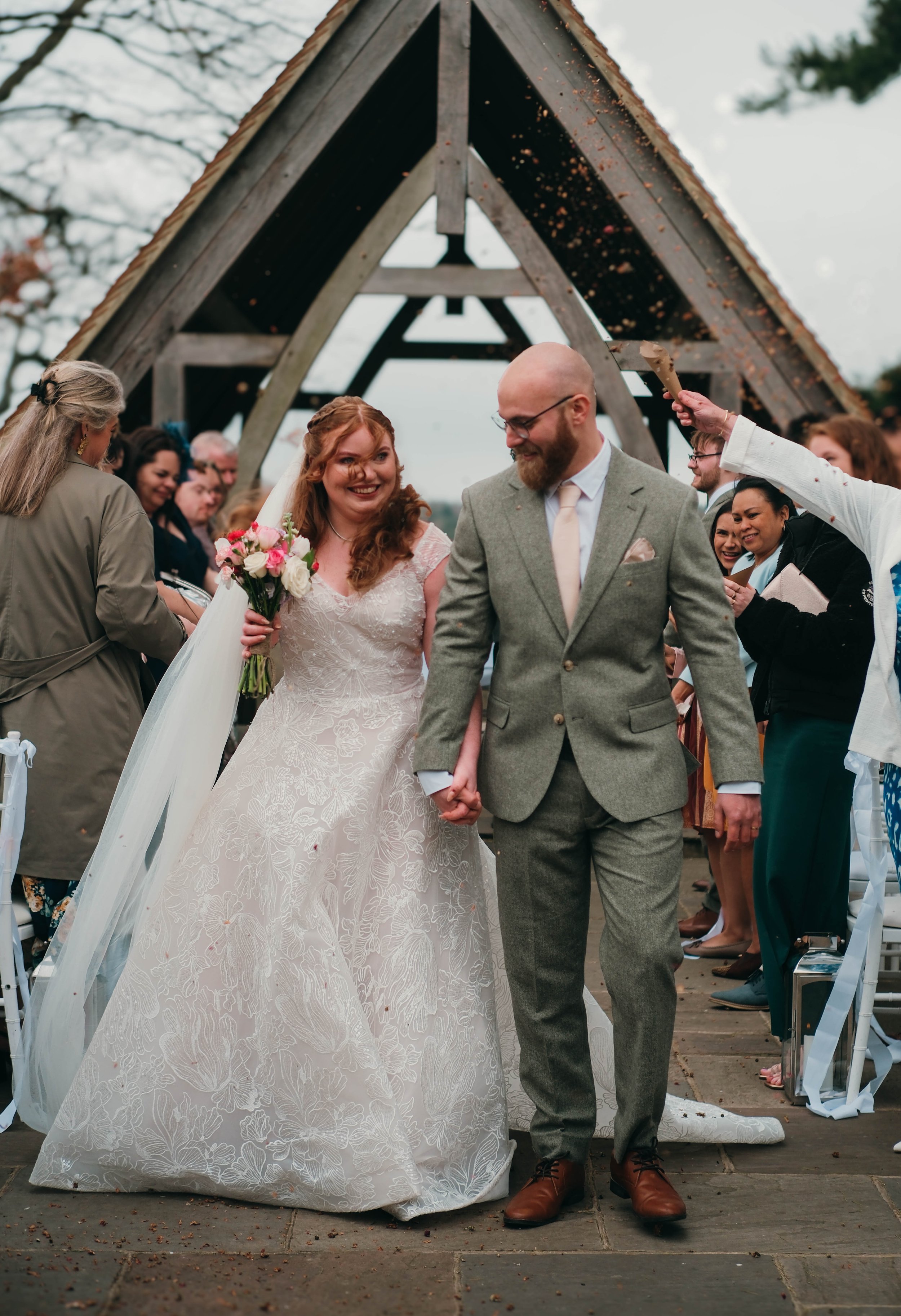 A wedding ceremony outdoors with the bride and groom holding hands under an A-shaped wooden arch. The bride is wearing a lace wedding gown and holding a bouquet of pink and white flowers, while the groom is dressed in a gray suit with a light pink ti