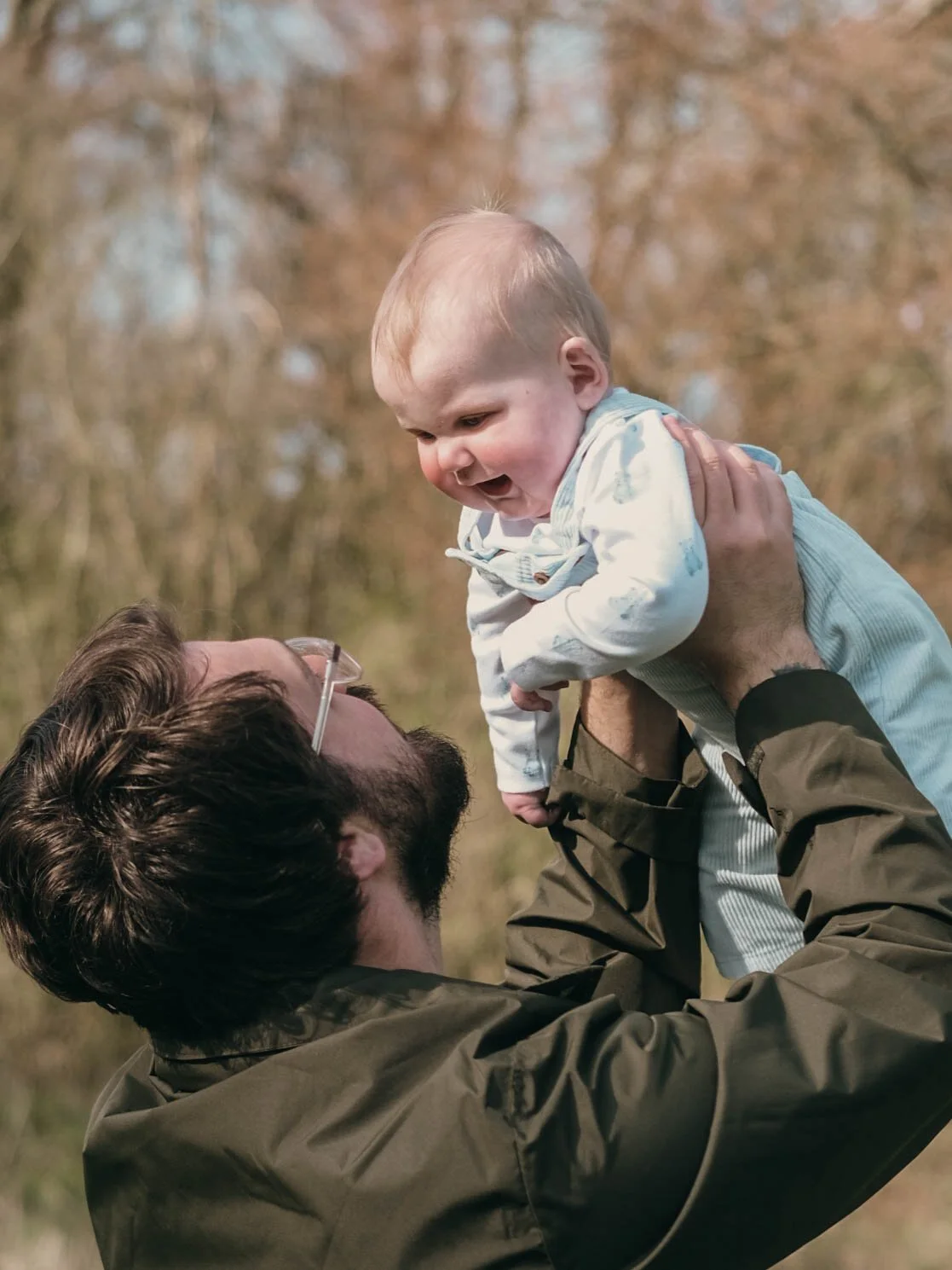 A man with glasses and a beard lifts a smiling baby boy into the air outdoors with trees in the background.