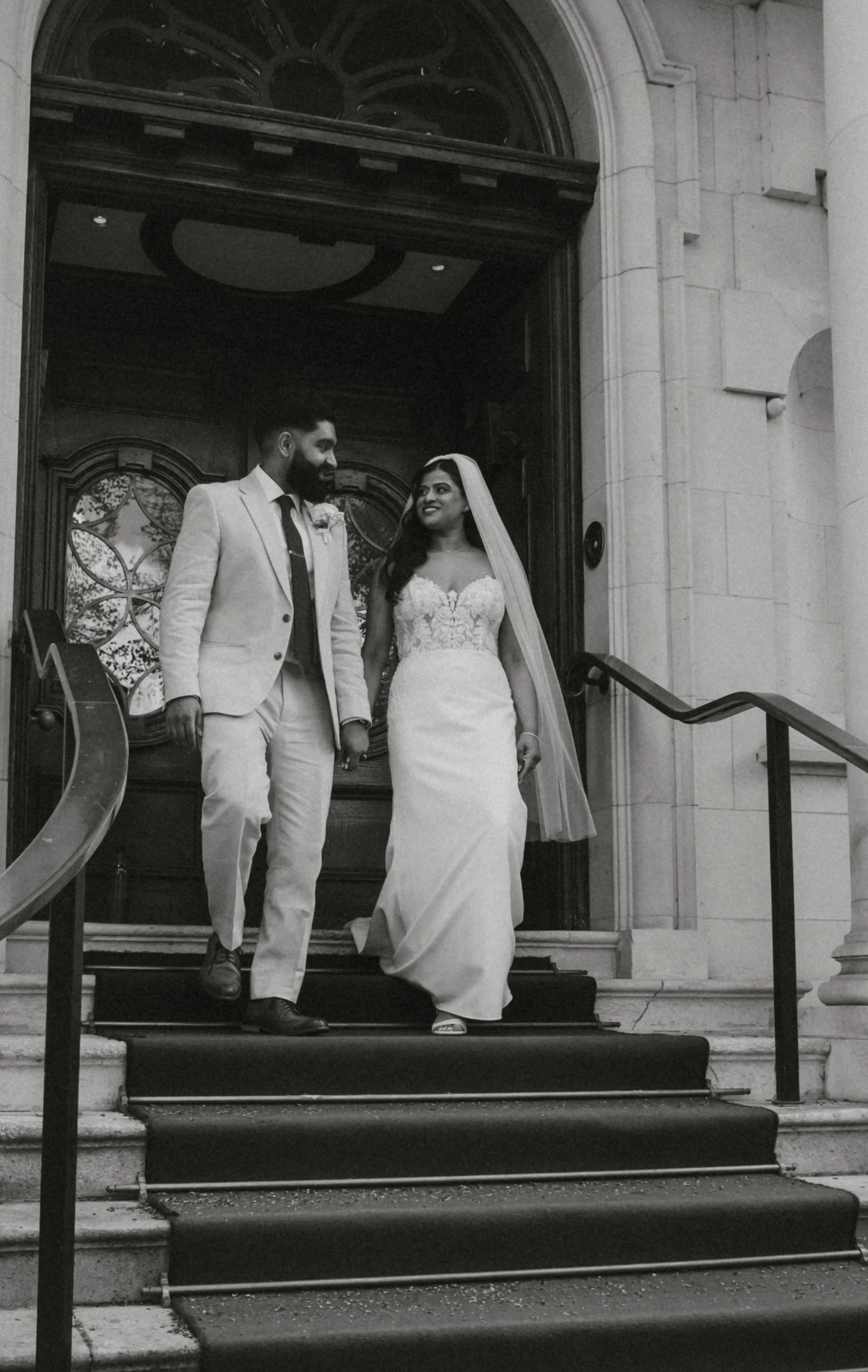 A bride and groom are walking down the steps of a church after their wedding, holding hands and looking at each other with smiles. The bride is wearing a white lace wedding gown with a veil, and the groom is dressed in a light-colored suit and tie.