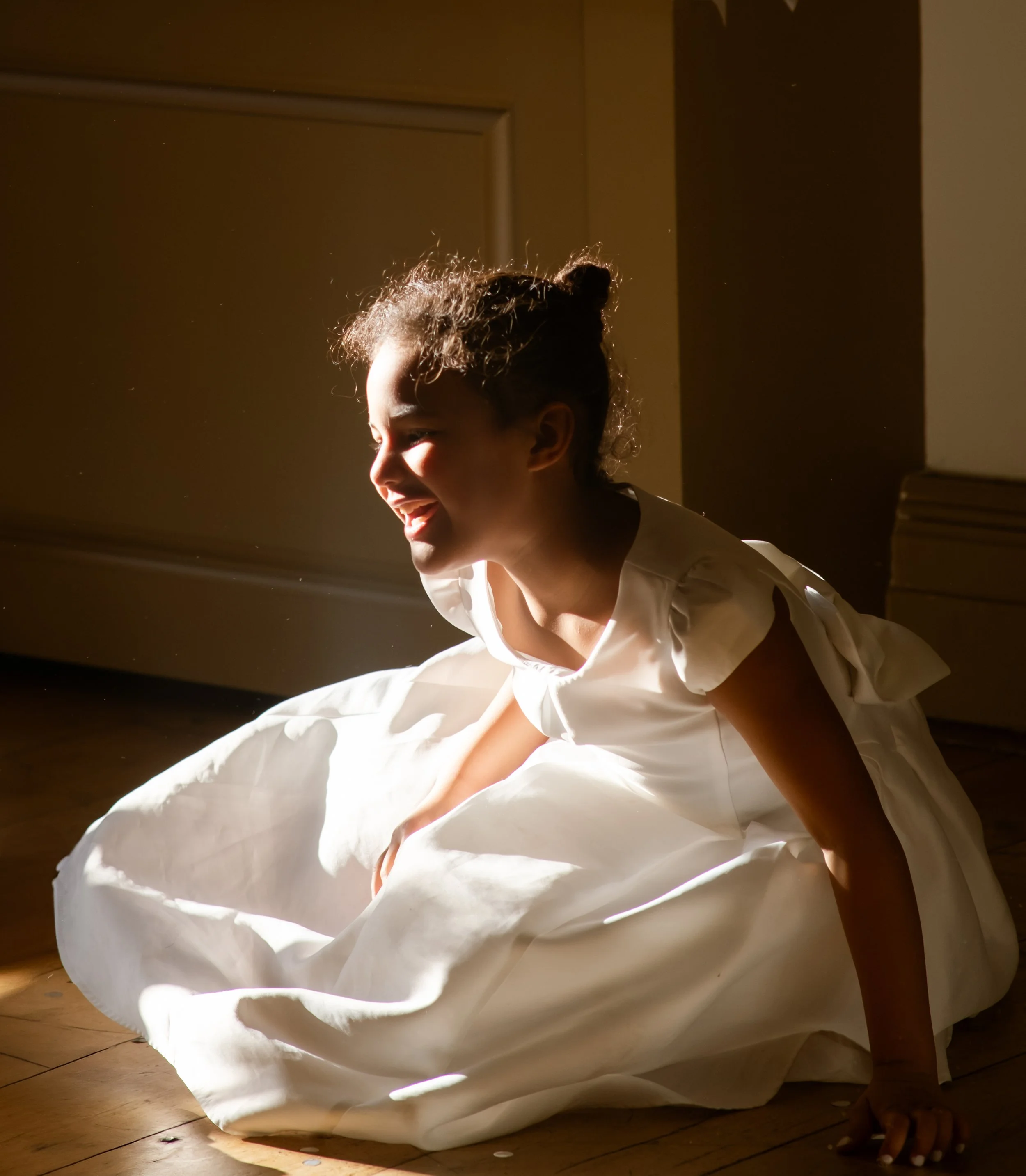 A woman with curly hair in a bun, smiling and wearing a white dress, sitting on a wooden floor with sunlight shining on her face, surrounded by falling confetti.