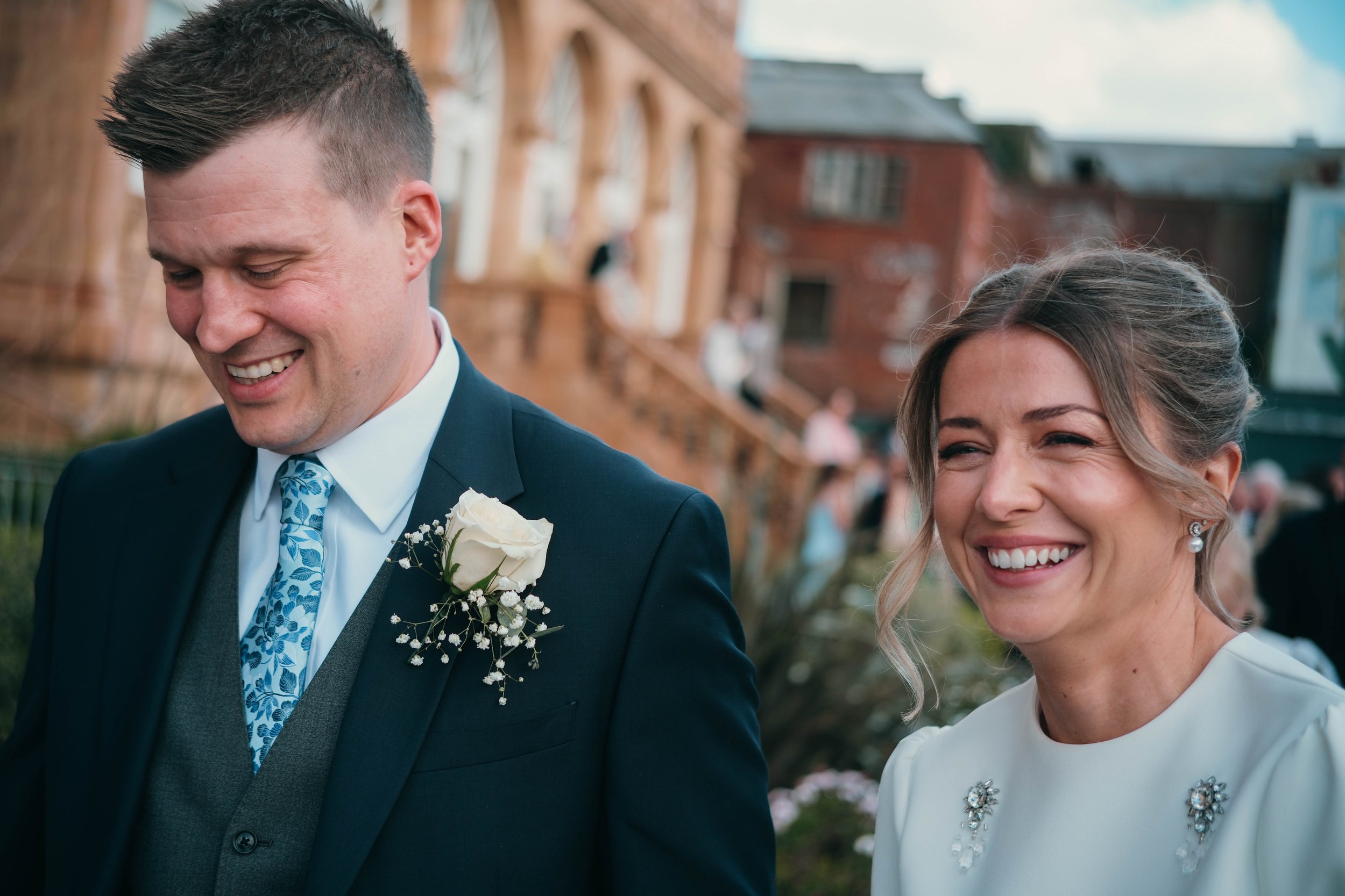A smiling couple at a wedding, the man with short dark hair wearing a dark suit, light blue tie, and boutonnière, and the woman with blonde hair in an elegant white dress with floral embellishments, standing outdoors.