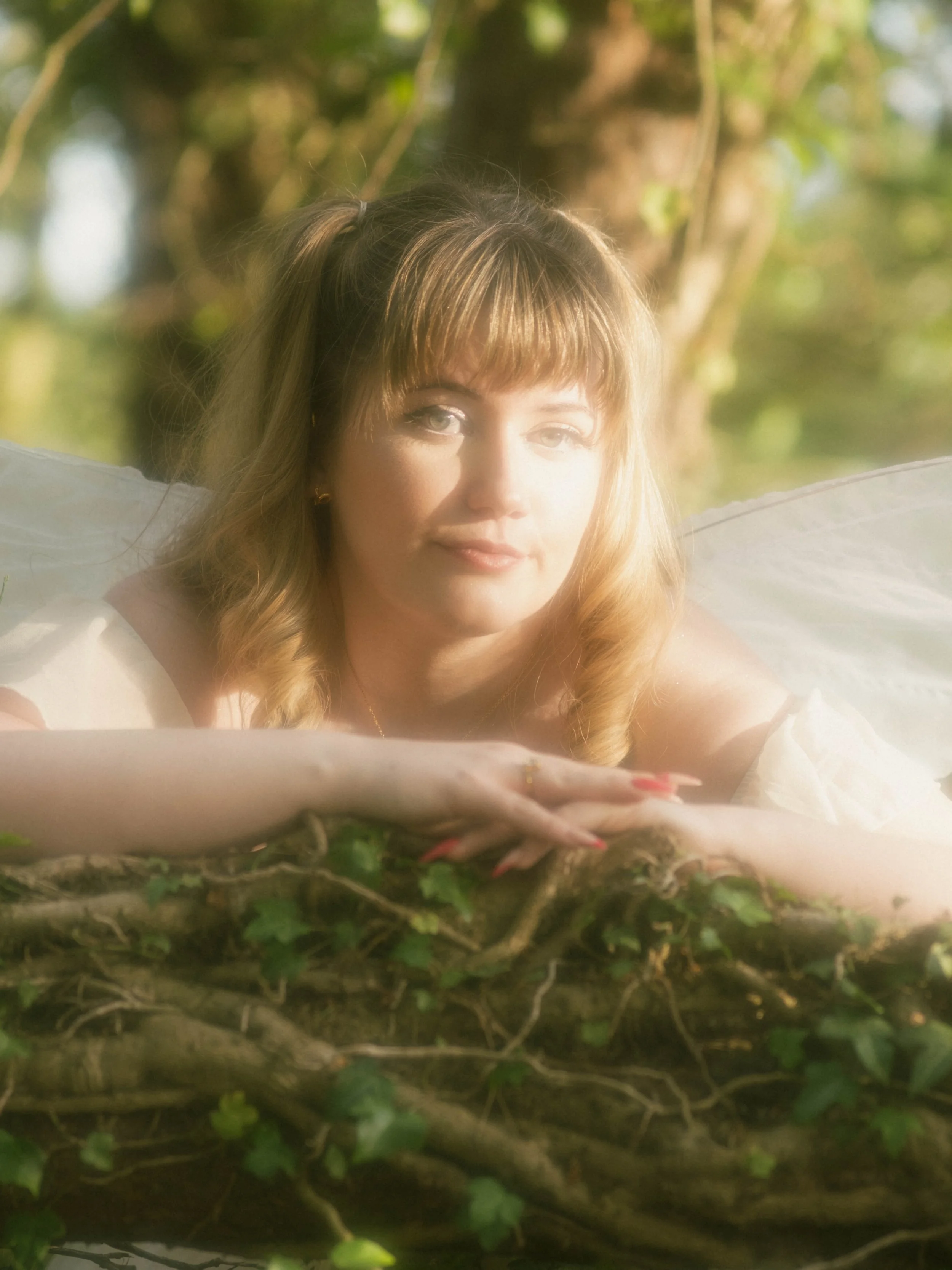 A woman with blonde hair and fairy wings lying on ground among greenery, with sunlight shining on her face.