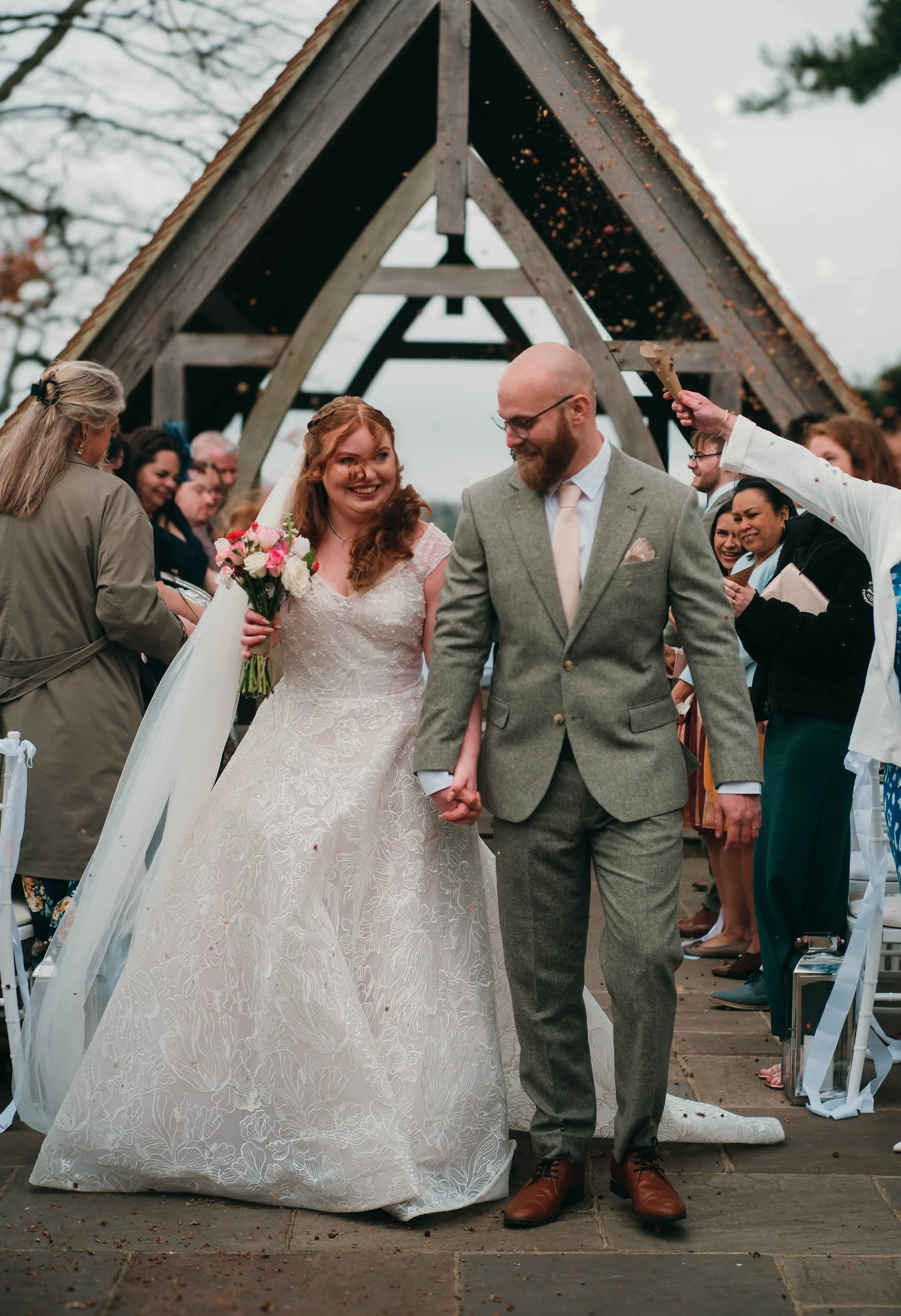 A couple gets married outdoors under a wooden A-frame structure, with a group of friends and family around them. The bride is holding a bouquet of pink and white flowers, and the groom is dressed in a light gray suit and tie. A person in white is using a small hammer to ring a bell above the couple.