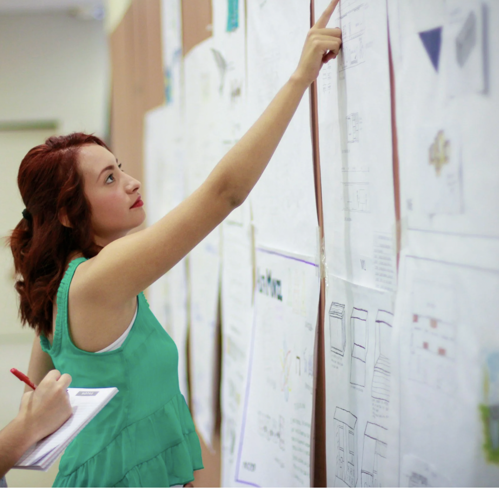 A woman with red hair wearing a green dress is pointing at a large whiteboard covered with paper diagrams and notes. She is holding a red pen and a notepad, and appears to be explaining or reviewing the information on the wall.