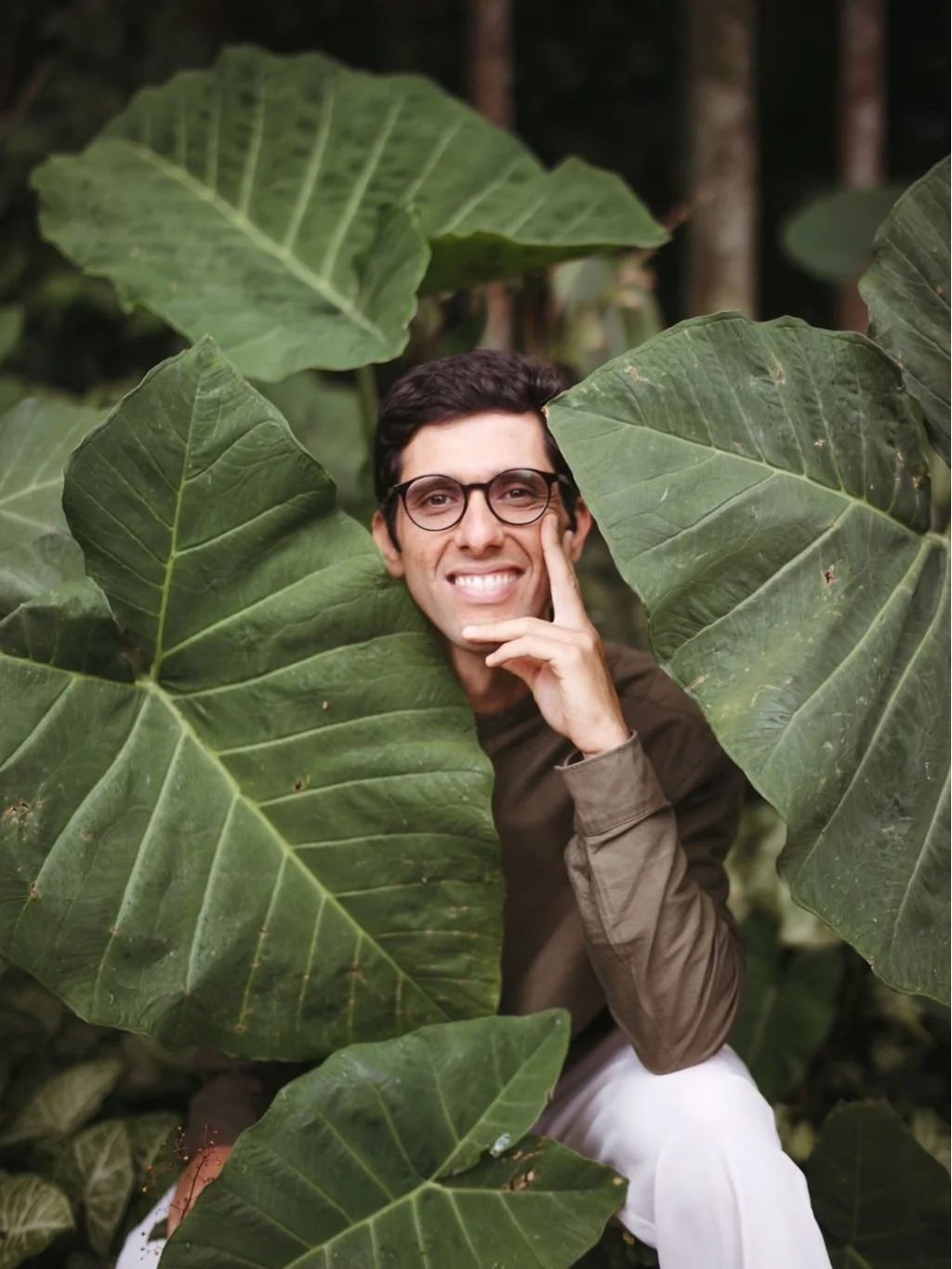 Homem sorridente usando óculos, cercado por grandes folhas verdes de plantas tropicais.