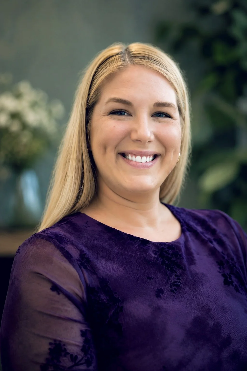 A woman with long blonde hair smiling, wearing a purple top, with a blurred green background.