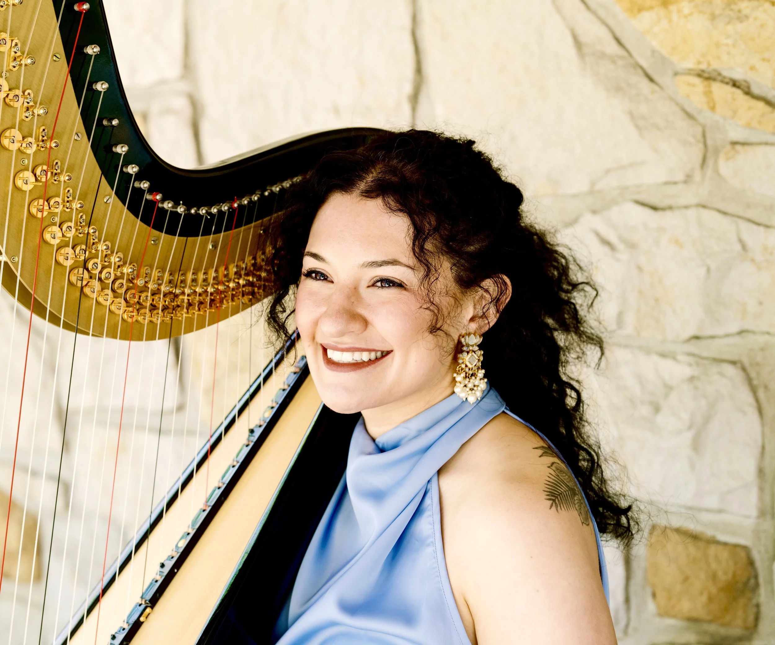 Hannah Anderson, harpist, is looking at the camera. She has dark, curly hair and is smiling, wearing large, ornate earrings, and blue sleeveless dress, playing a harp with a stone wall in the background.