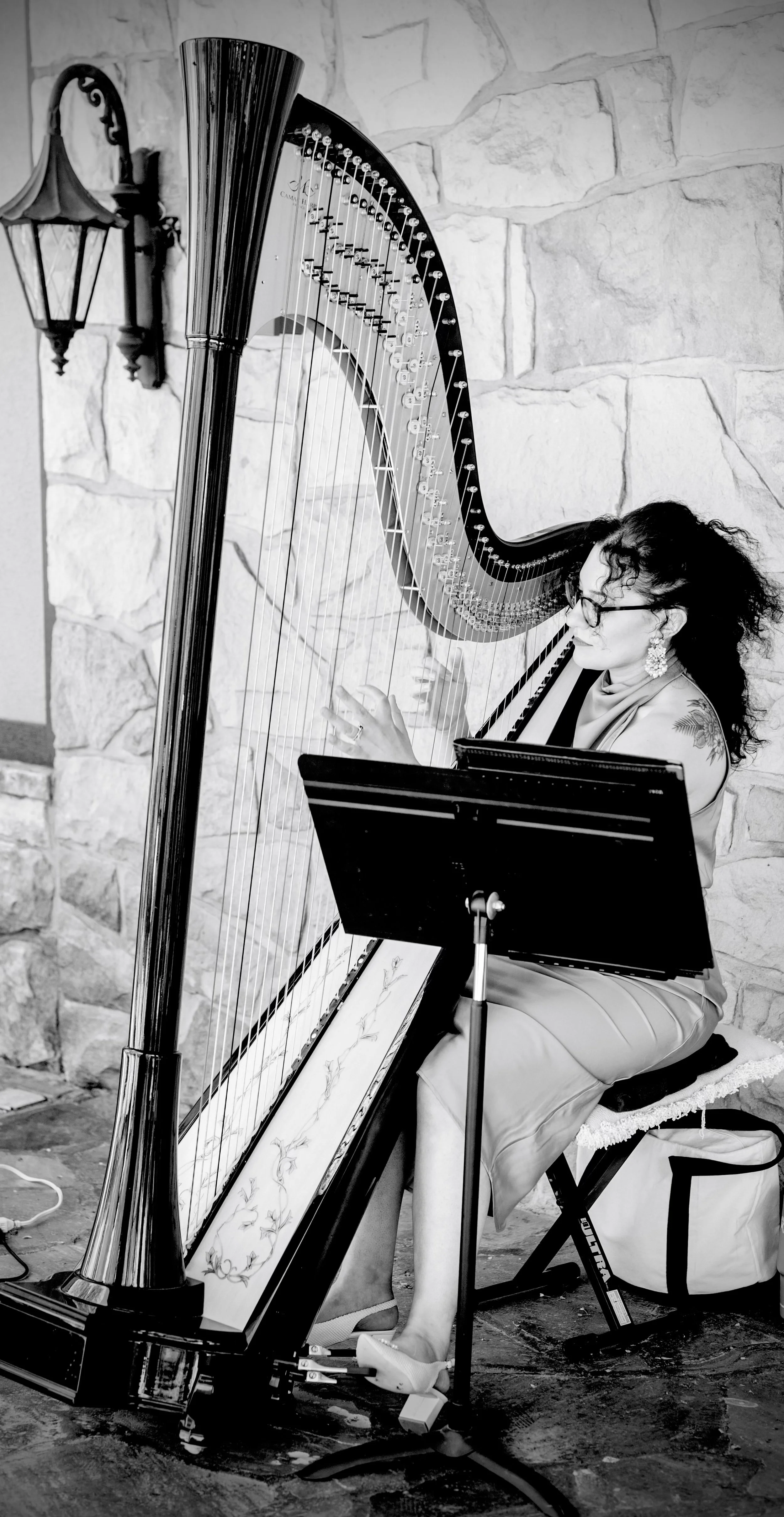 Woman playing a harp, sitting on a chair, with sheet music stand, against a stone wall backdrop