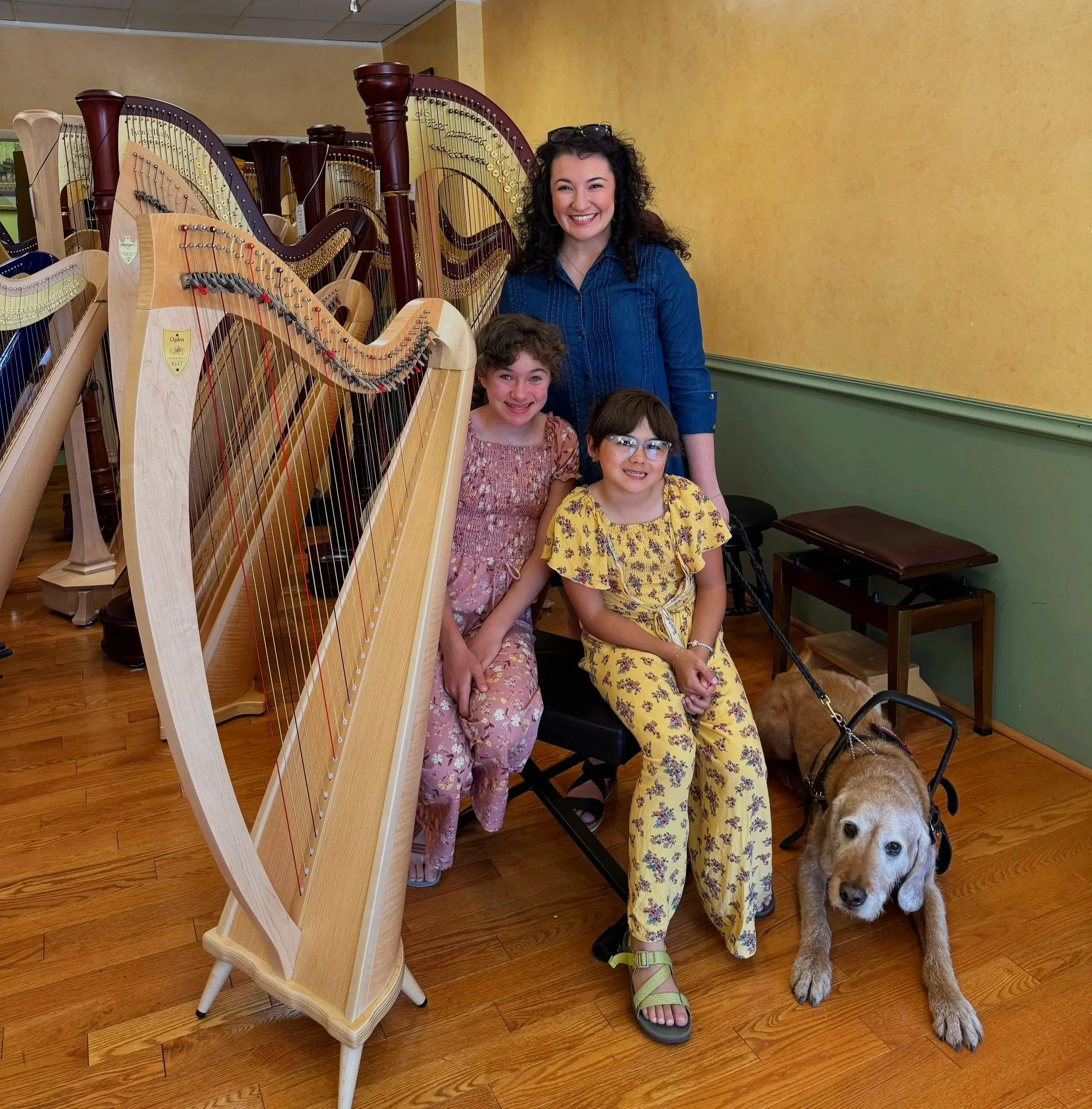 A woman and two children smiling, standing next to a large wooden harp, with a guide dog next to them, in an indoor room with yellow and green walls and wooden flooring.