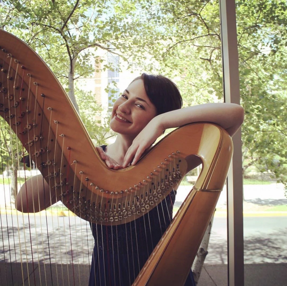 Hannah Anderson, harpist, circa 2015. Hannah stands behind a blonde harp, her arms are on top of the harp. She is looking at the camera and smiling